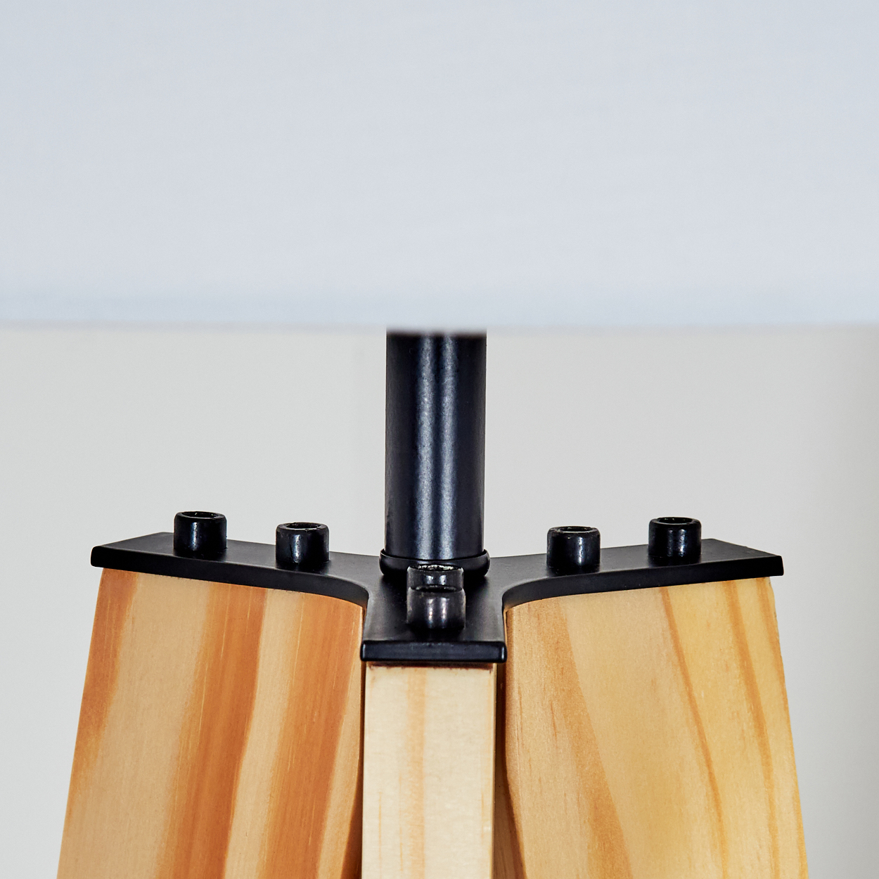Nahaufnahme eines Lampenfußes. Er hat Holzbeine, schwarze Metallstützen und eine schwarze Stange. Oben sitzt ein weißer Lampenschirm.