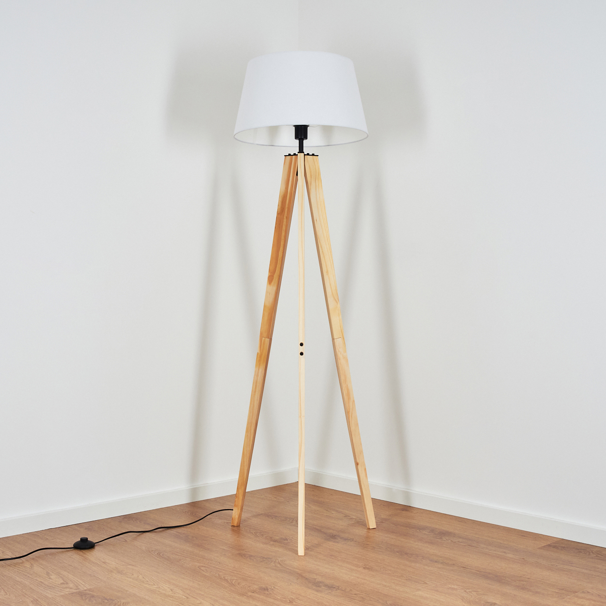 Eine hölzerne Stativ-Stehlampe mit weißem Lampenschirm steht in einer Ecke vor einer weißen Wand und einem Holzboden.