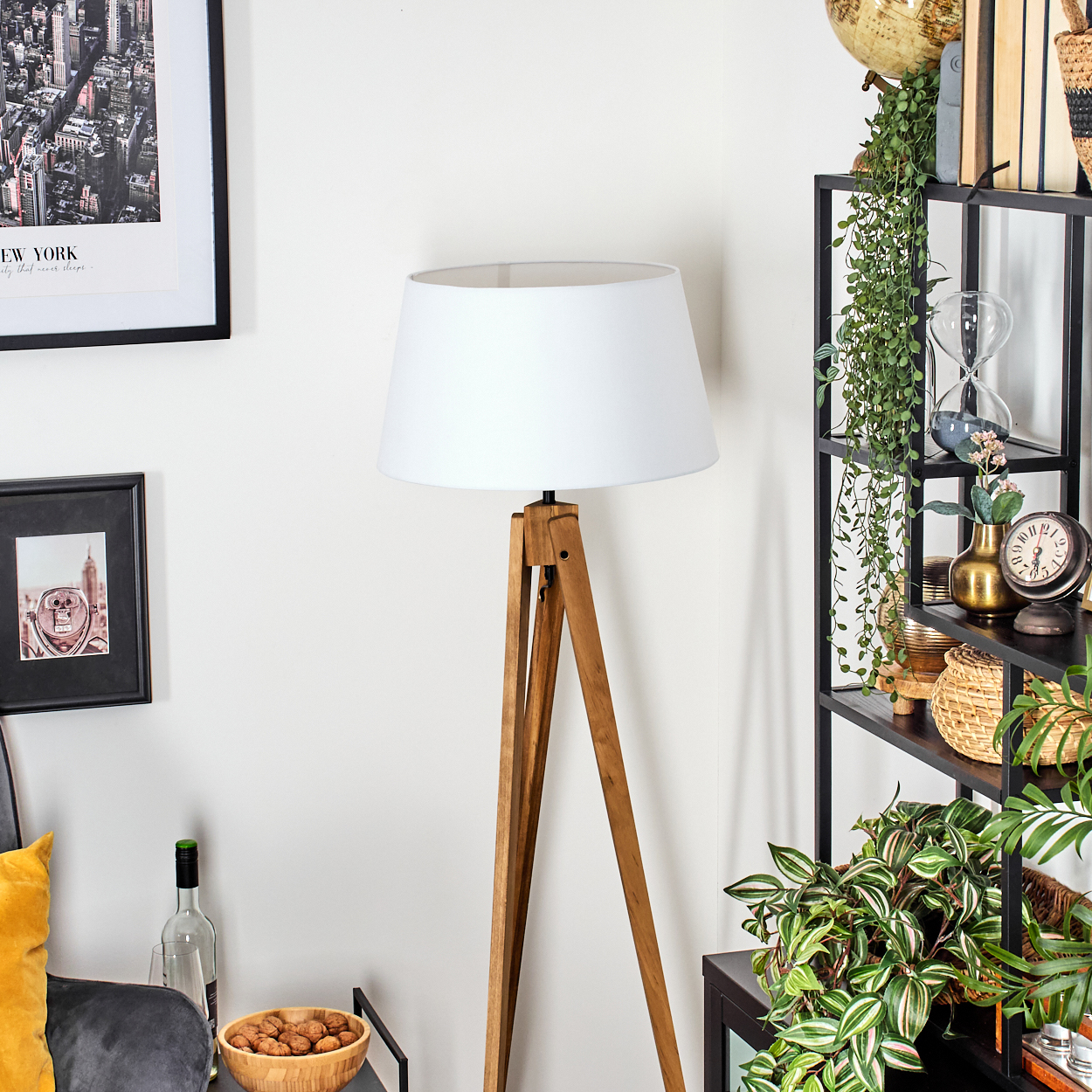 Eine Holz-Stativ-Stehlampe mit weißem Lampenschirm steht neben einem schwarzen Regal mit Pflanzen und Dekorationsartikeln.