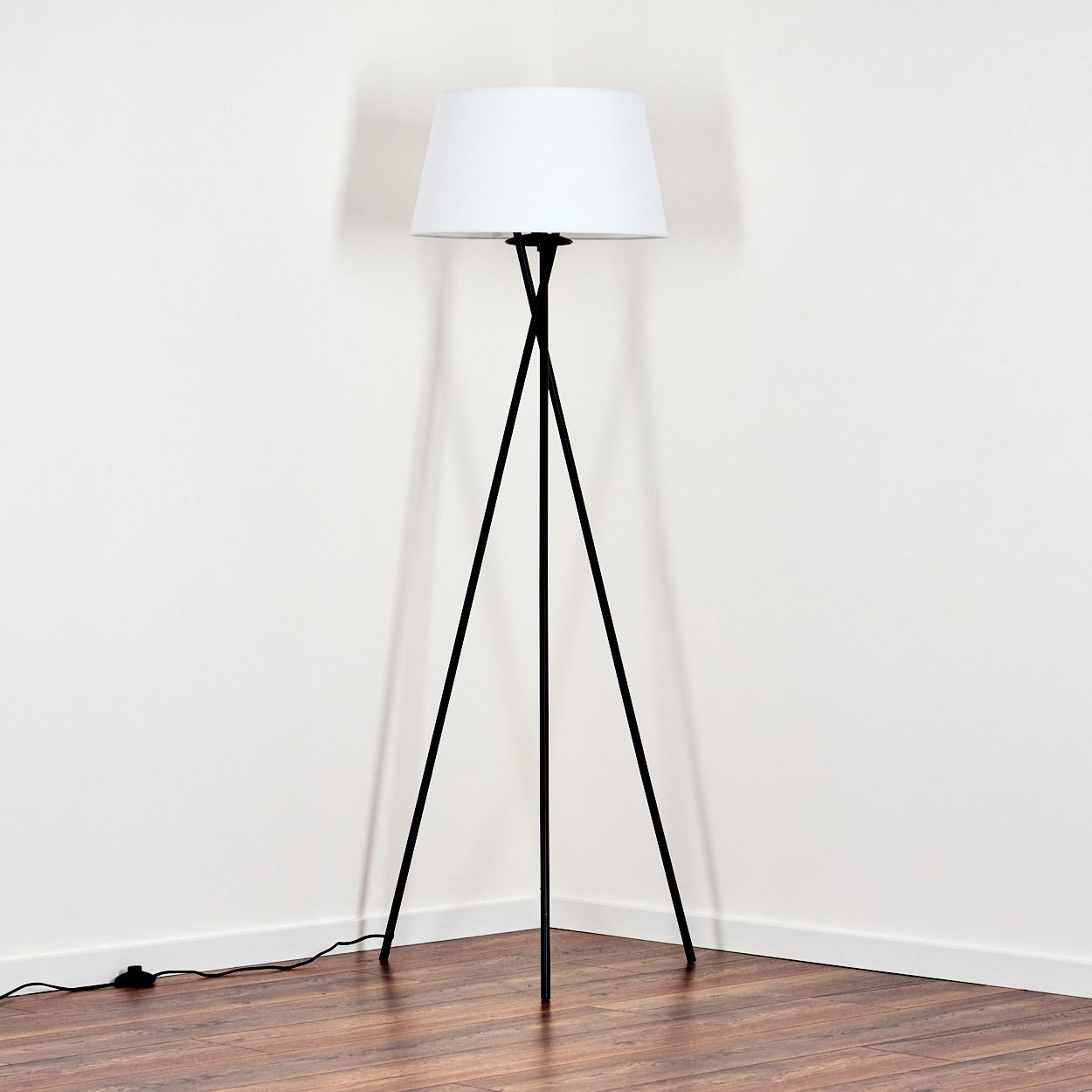 Eine schwarze Stehlampe mit weißem Lampenschirm steht in einer Ecke. Der Boden ist aus Holz, die Wände sind weiß.