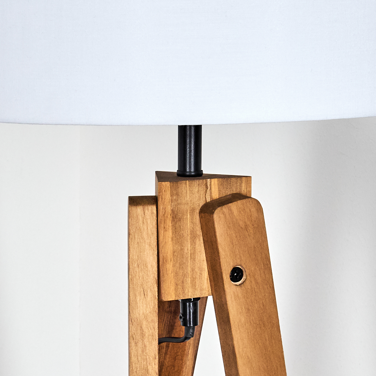 Ein Lampenstativ aus Holz trägt einen weißen Lampenschirm. Der Sockel hat eine schwarze Metallkomponente. Der Hintergrund ist eine schlichte weiße Wand.