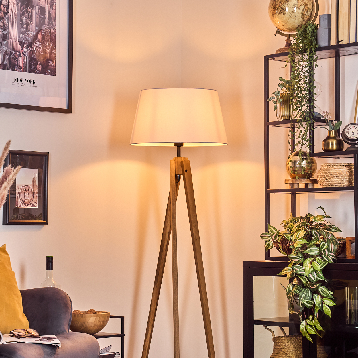 Ein Raum mit einer hölzernen Stativ-Stehlampe. Regale mit Pflanzen und Dekoration sind zu sehen. Ein gerahmtes New York-Poster und ein Sofa sind im Bild.