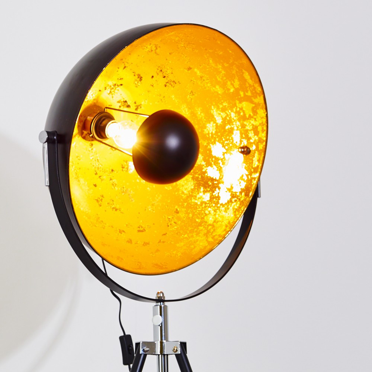 Eine schwarze und goldene Stehlampe leuchtet vor einem weißen Hintergrund. Sie hat ein Stativ und einen großen, runden Schirm.