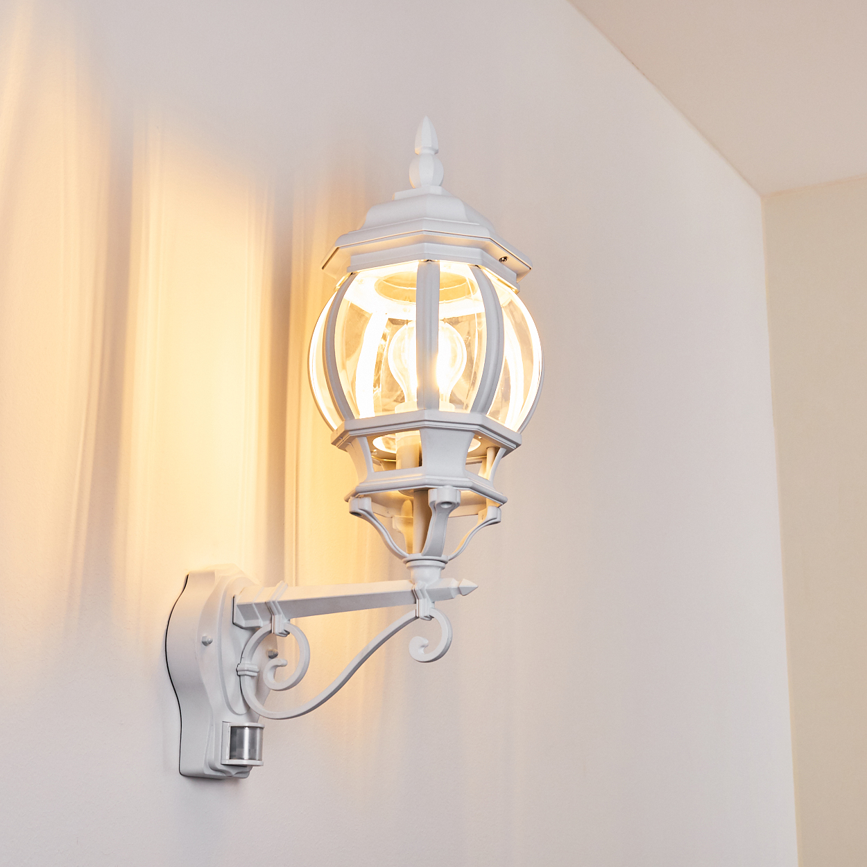 Weiße Außenlampe an einer weißen Wand. Die Lampe ist beleuchtet, mit Glaskugel und Verzierungen. Die Szene ist gut beleuchtet.