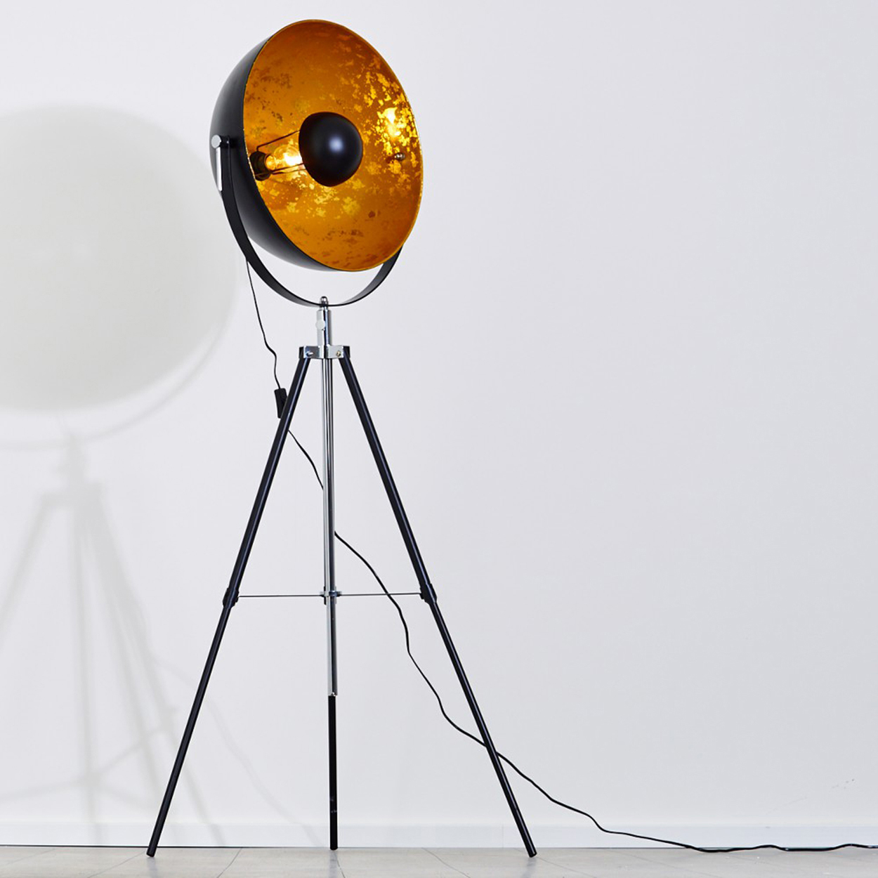 Eine schwarz-goldene Stehlampe mit Stativfuß, die einen Schatten auf eine weiße Wand wirft. Die Lampe hat einen großen, runden Schirm.
