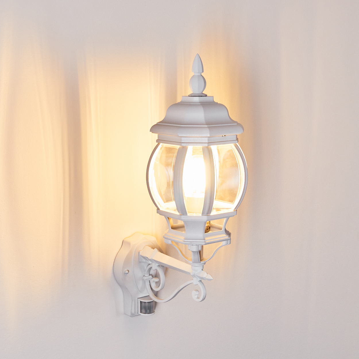 Eine weiße Außenlampe ist an einer Wand befestigt. Die Lampe hat ein kugelförmiges Glasgehäuse und ist beleuchtet. Die Wand ist weiß.