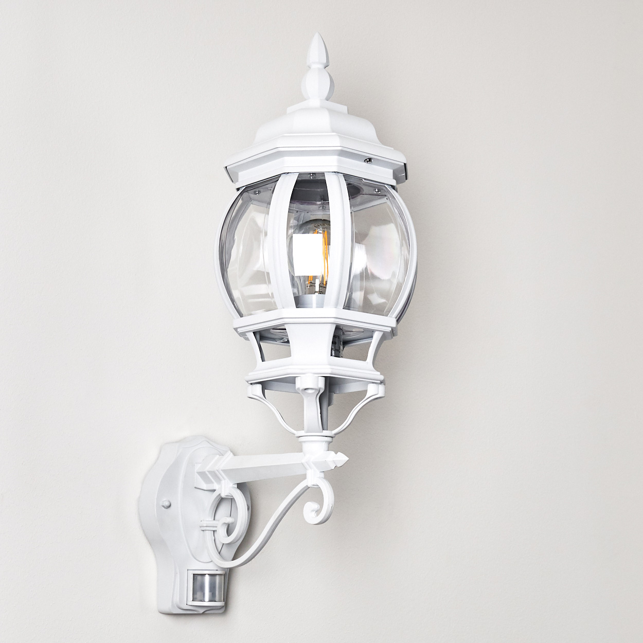 Eine weiße Außenlampe ist an einer Wand montiert. Sie hat ein dekoratives Design, klares Glas und eine Glühbirne. Ein Bewegungssensor ist vorhanden.