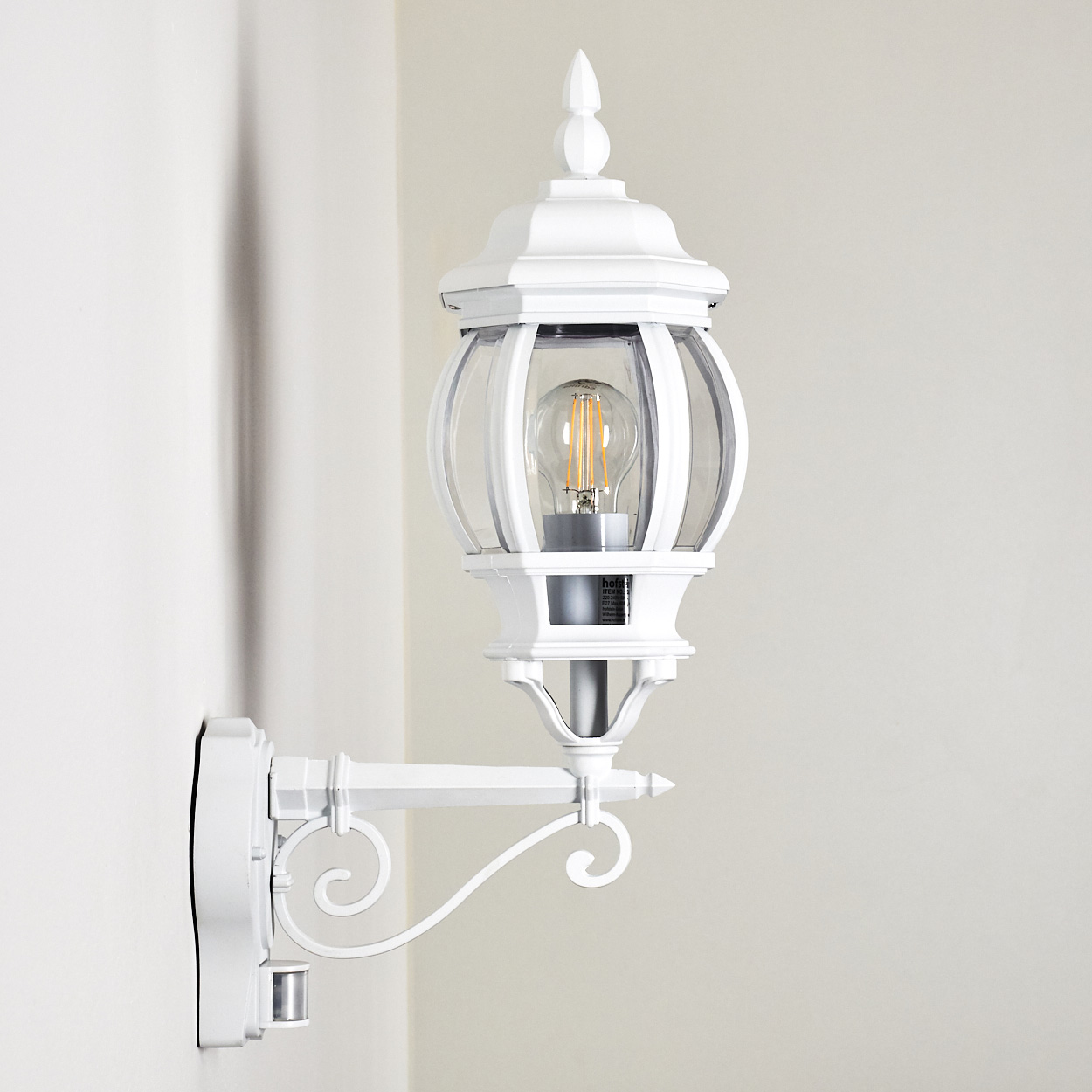 Weiße Außenlampe an einer weißen Wand. Sie hat einen Glaskolben und eine LED-Lampe. Die Lampe ist an einem gebogenen Arm montiert.