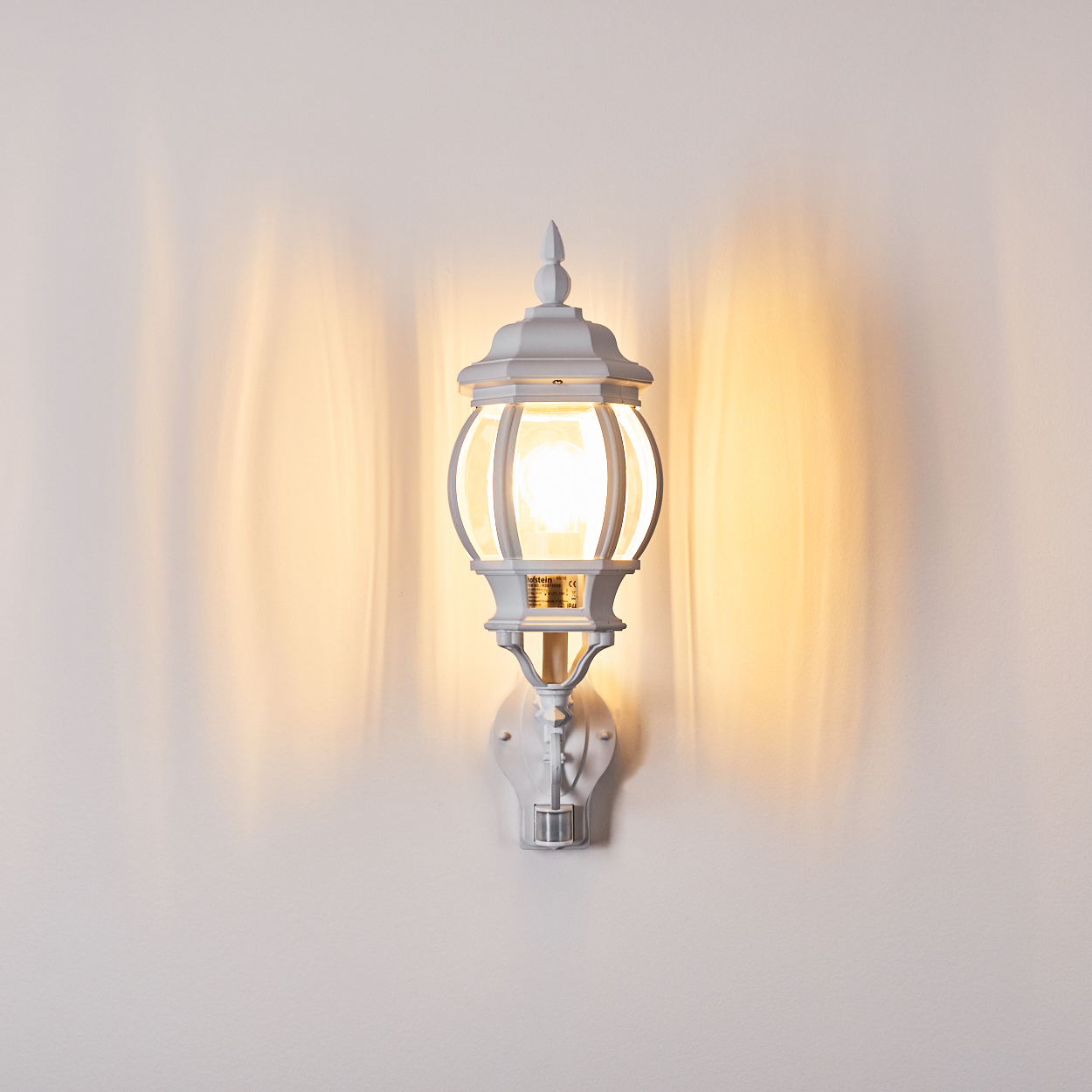 Eine weiße Außenlampe ist an einer Wand befestigt. Die Lampe hat einen Glasschirm und ist beleuchtet. Die Wand ist hell.