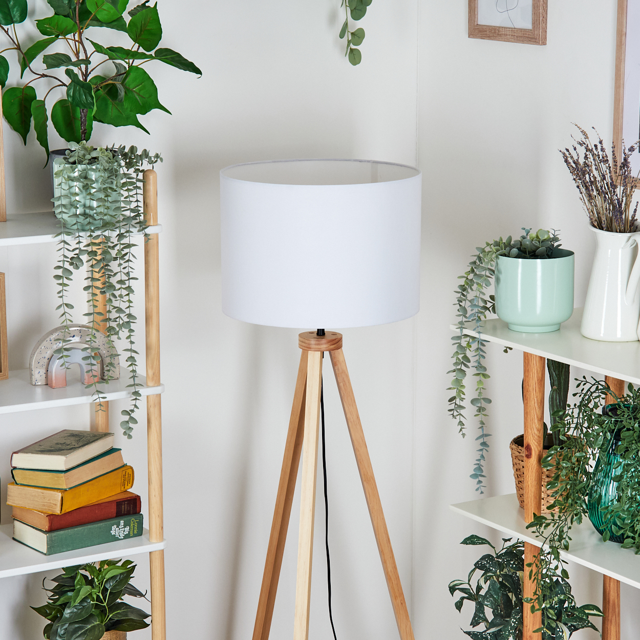 Eine weiße Stehlampe mit Holzstativ steht neben Regalen mit Pflanzen, Büchern und Dekoration. Ein heller Raum.