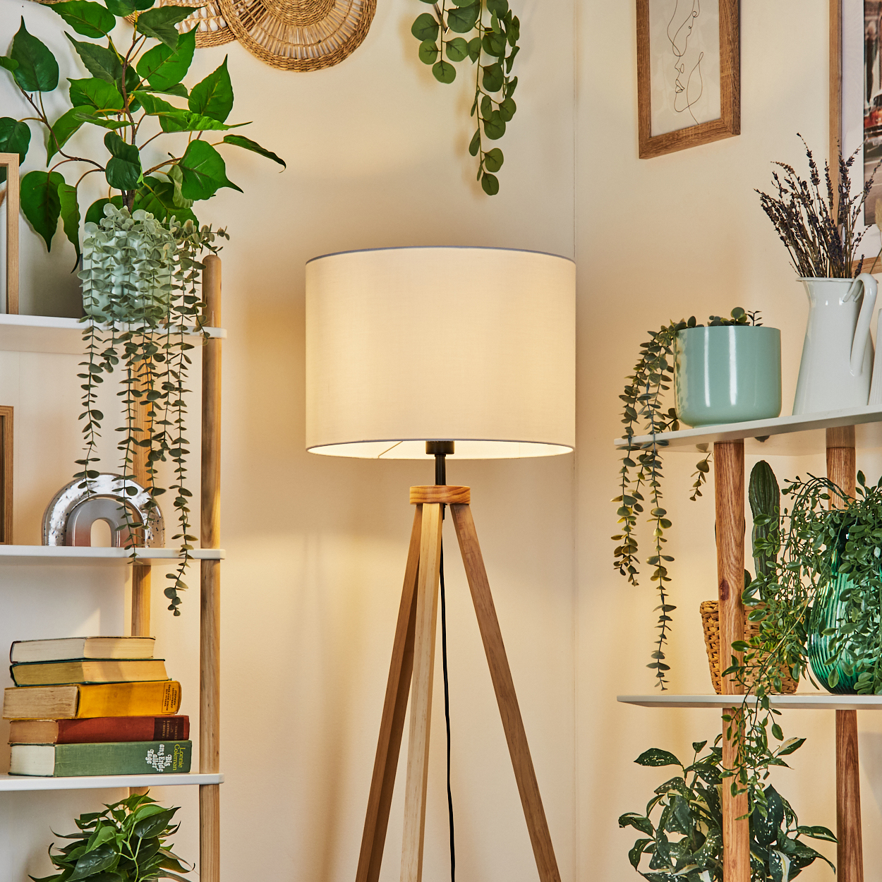 Eine hölzerne Stativlampe steht in einem Raum mit Regalen, Pflanzen und Büchern. Ein weißer Lampenschirm befindet sich oben. Die Wand ist beige.