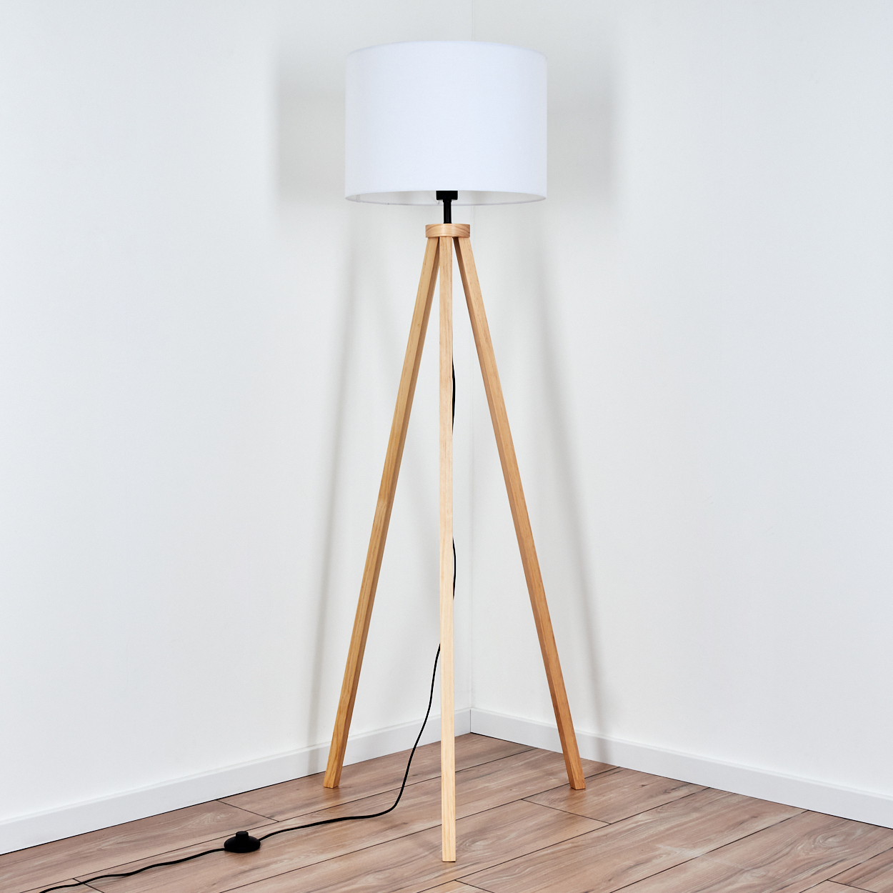 Eine hölzerne Stativ-Stehlampe mit weißem Lampenschirm steht in der Ecke eines Raumes. Der Boden ist aus Holz, die Wände sind weiß.