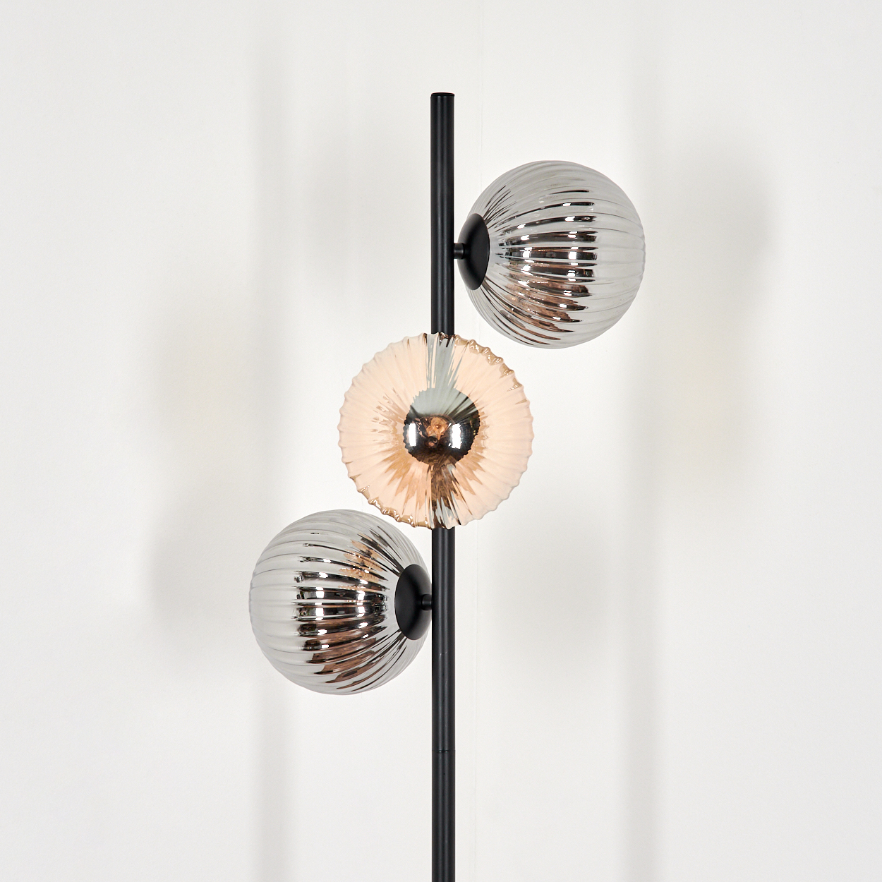 Eine moderne Lampe mit drei Leuchten. Zwei sind gerippte Kugeln, eine ist eine geriffelte Scheibe. Alle sind an einer schwarzen Stange montiert.