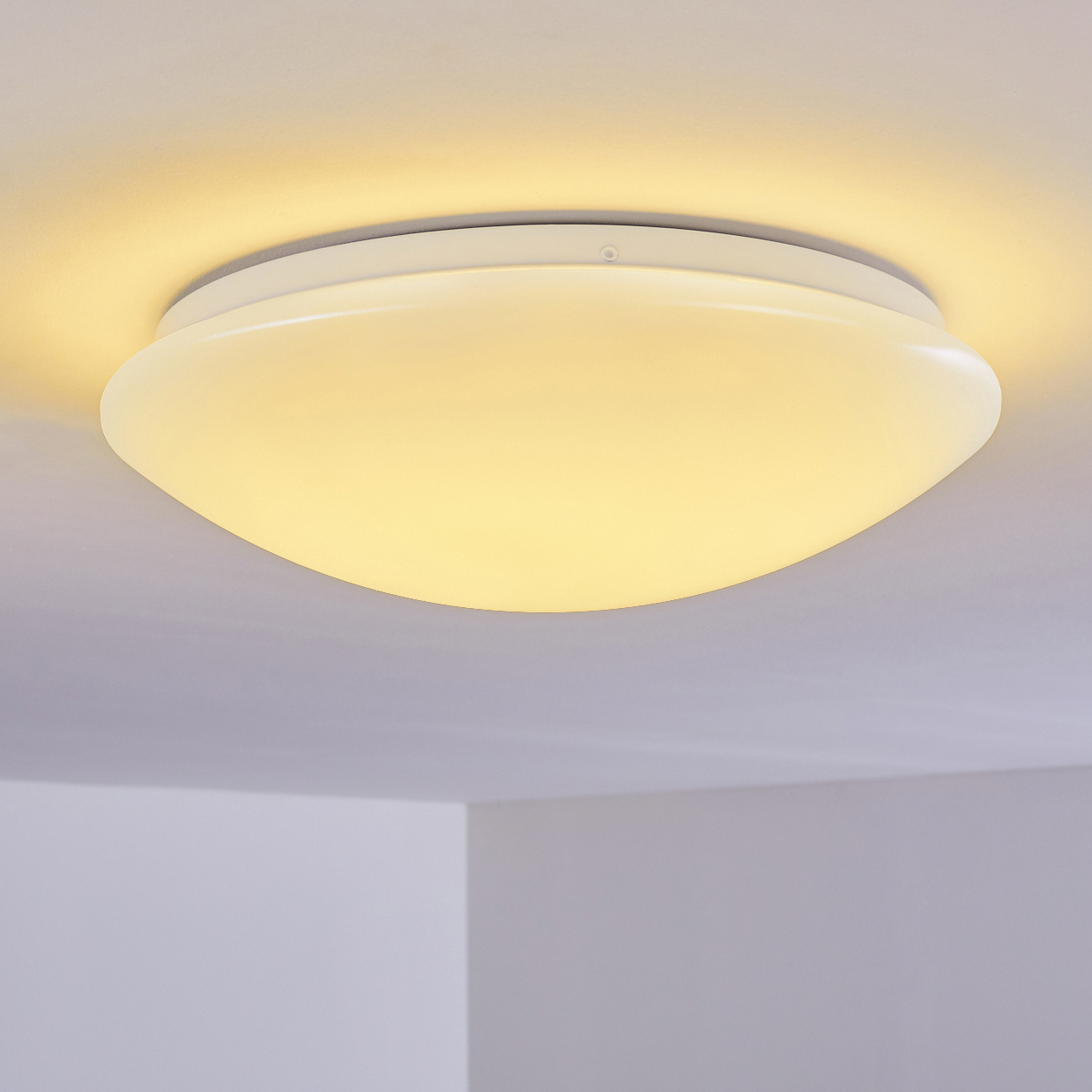 Eine weiße, abgerundete Deckenleuchte ist mittig im Bild. Sie strahlt ein warmes, gelbes Licht aus. Der Hintergrund zeigt eine weiße Decke.