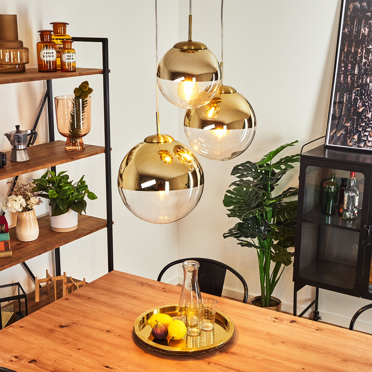 Ein Esszimmer mit Holztisch, goldenen Pendelleuchten und einem Tablett mit Obst. Regale und ein Schrank befinden sich im Hintergrund.