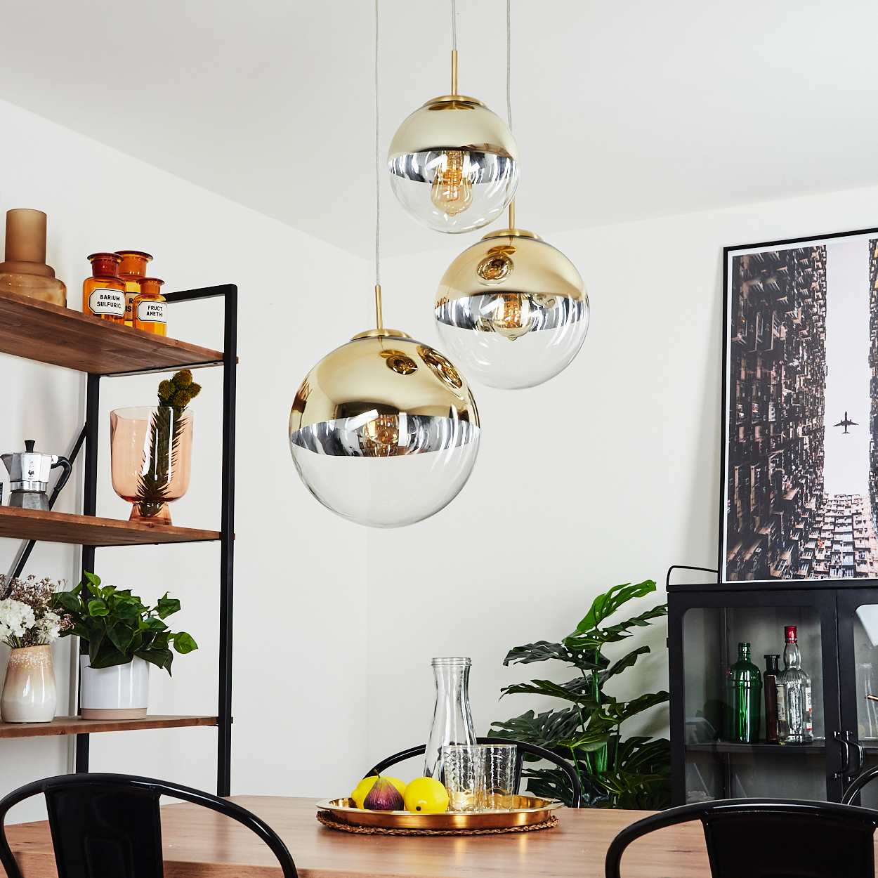 Ein Esszimmer mit hängenden Kugelleuchten. Ein Tisch ist mit Obst, Gläsern und einem Tablett gedeckt. Regale und ein gerahmtes Bild sind sichtbar.