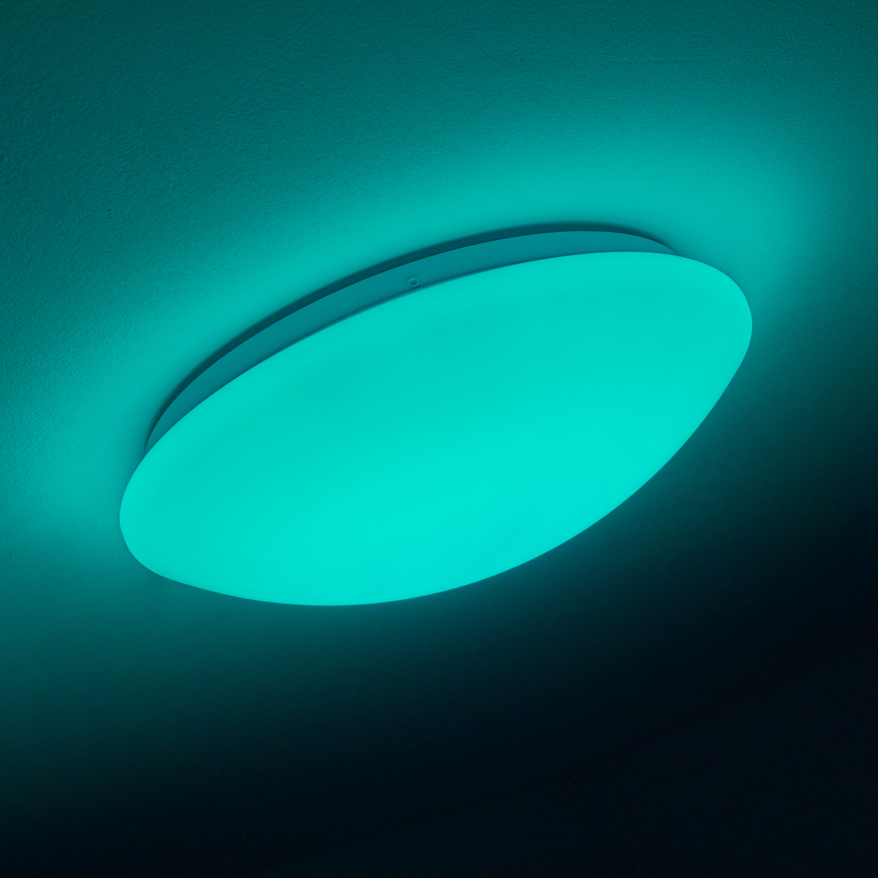 Eine runde, türkis farbene Deckenleuchte ist an einer weißen Decke montiert. Das Licht strahlt sanft türkis.