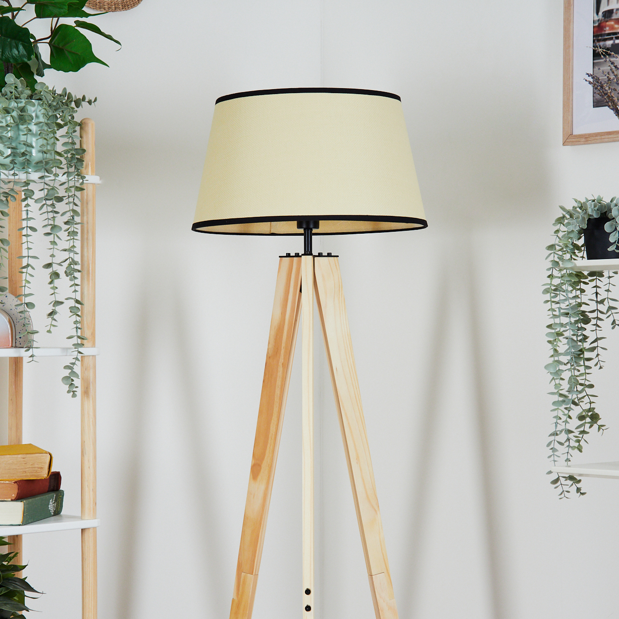 Eine hölzerne Stativlampe mit cremefarbenem Lampenschirm und schwarzer Verkleidung. Pflanzen und Regale sind im Hintergrund.