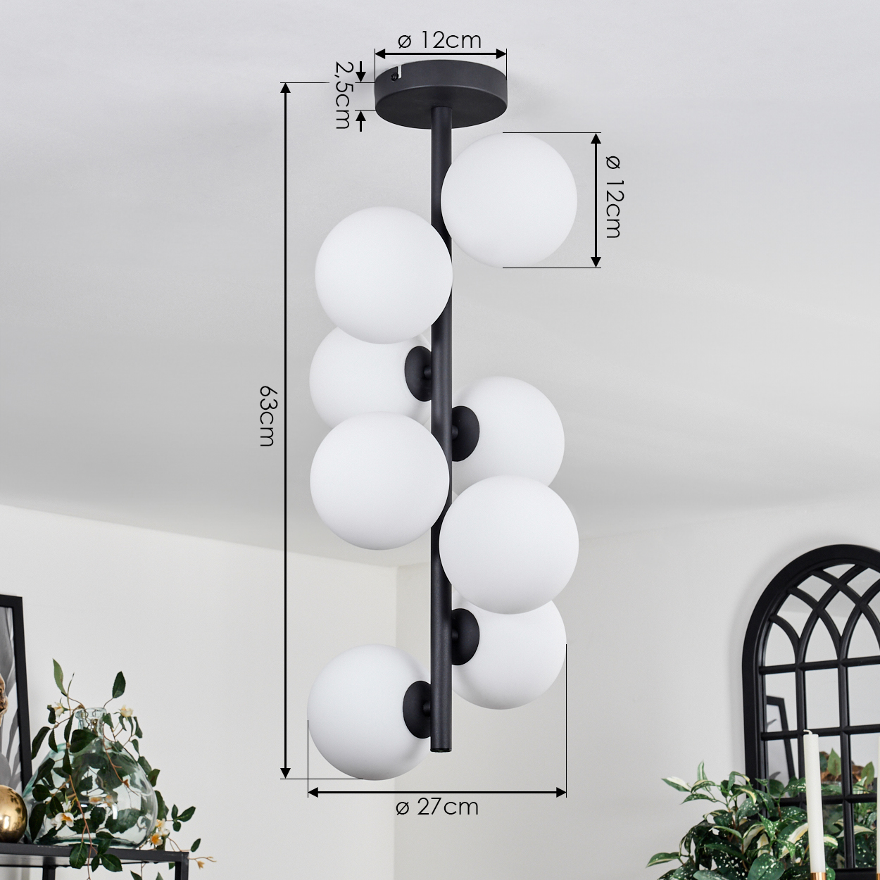 Eine schwarze moderne Deckenleuchte mit acht weißen kugelförmigen Lampen, Maße beschriftet. Hintergrund: weiße Decke und Pflanzen.