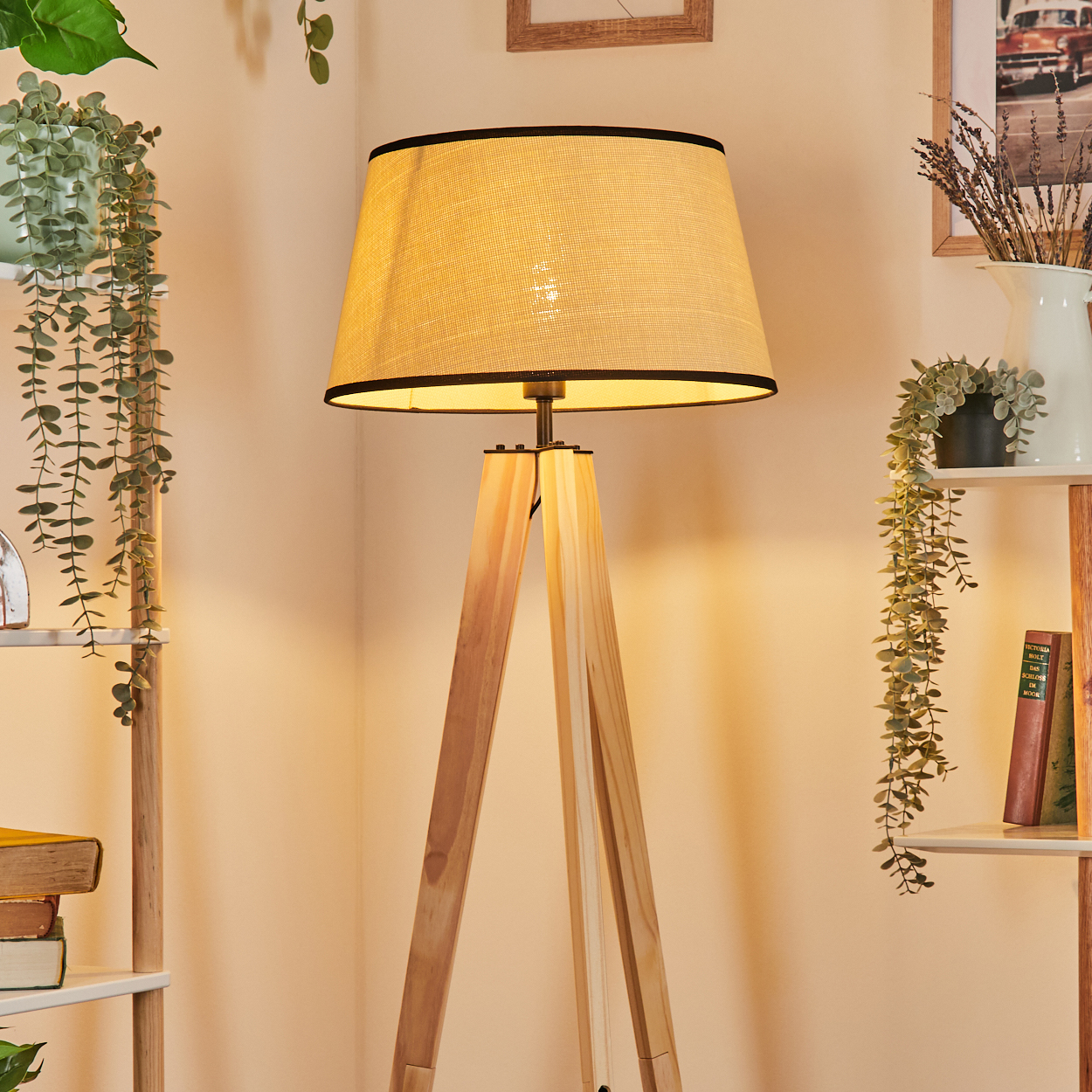 Eine hölzerne Stehlampe mit beigem Lampenschirm. Pflanzen und Regale mit Büchern schmücken den Hintergrund.
