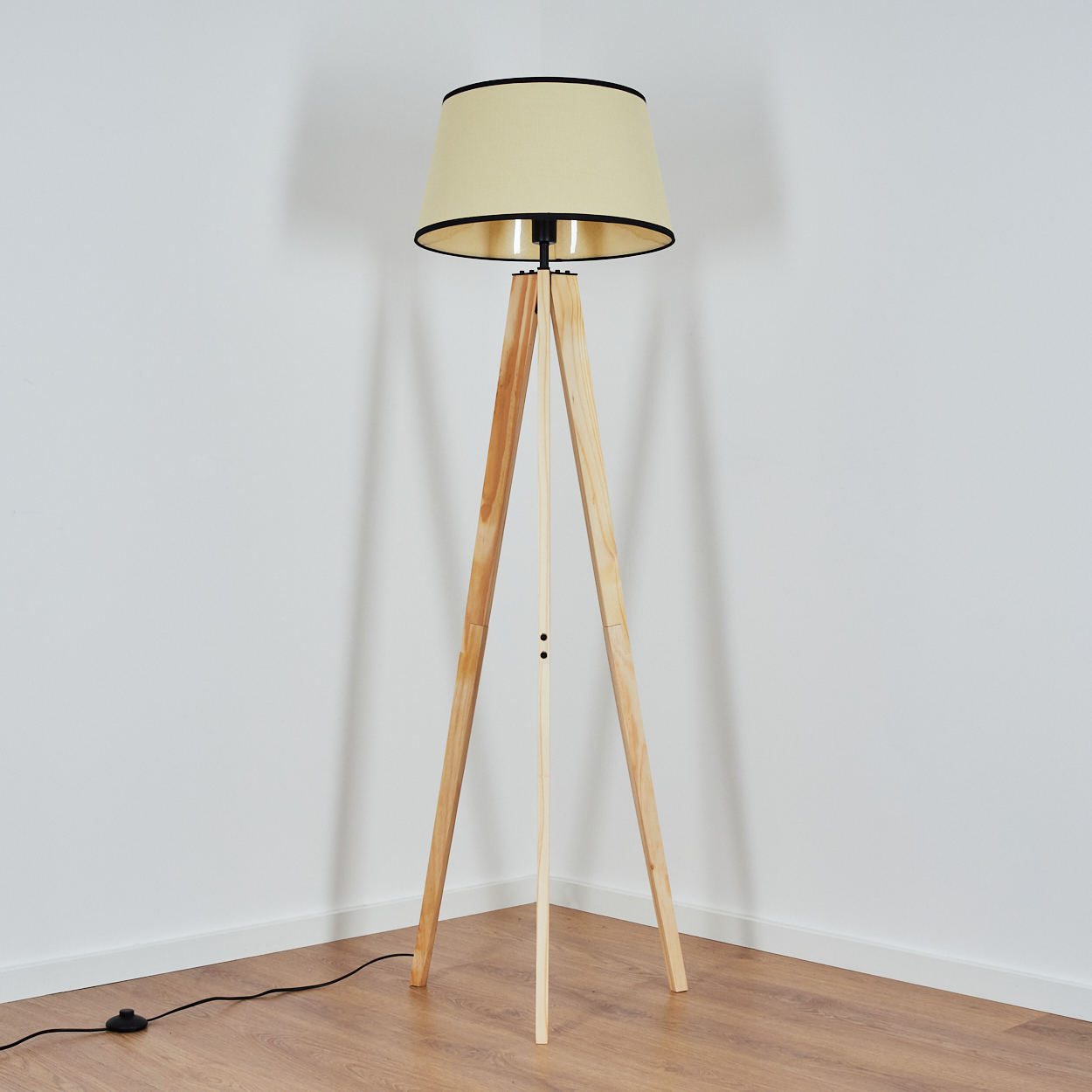Eine hölzerne Stativ-Stehlampe mit beigem Lampenschirm steht in einer Ecke. Der Boden ist aus Holz und die Wände sind weiß.