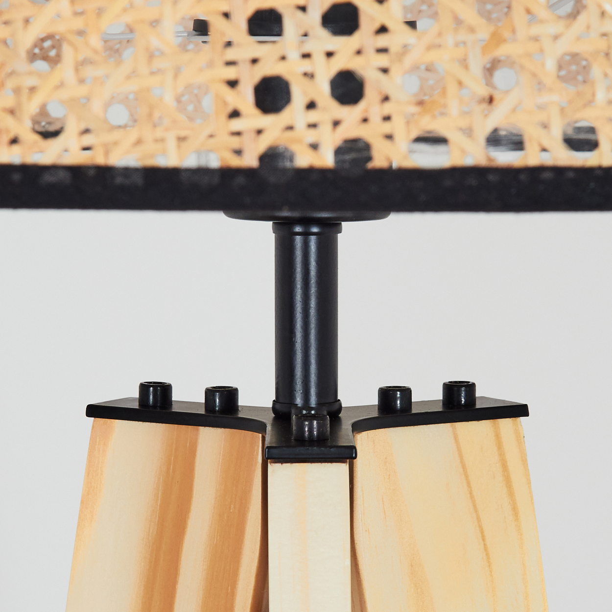 Nahaufnahme einer Lampe. Gewebter Lampenschirm über schwarzem Besatz, getragen von einer schwarzen Metallstange. Holzsockel mit Schrauben.