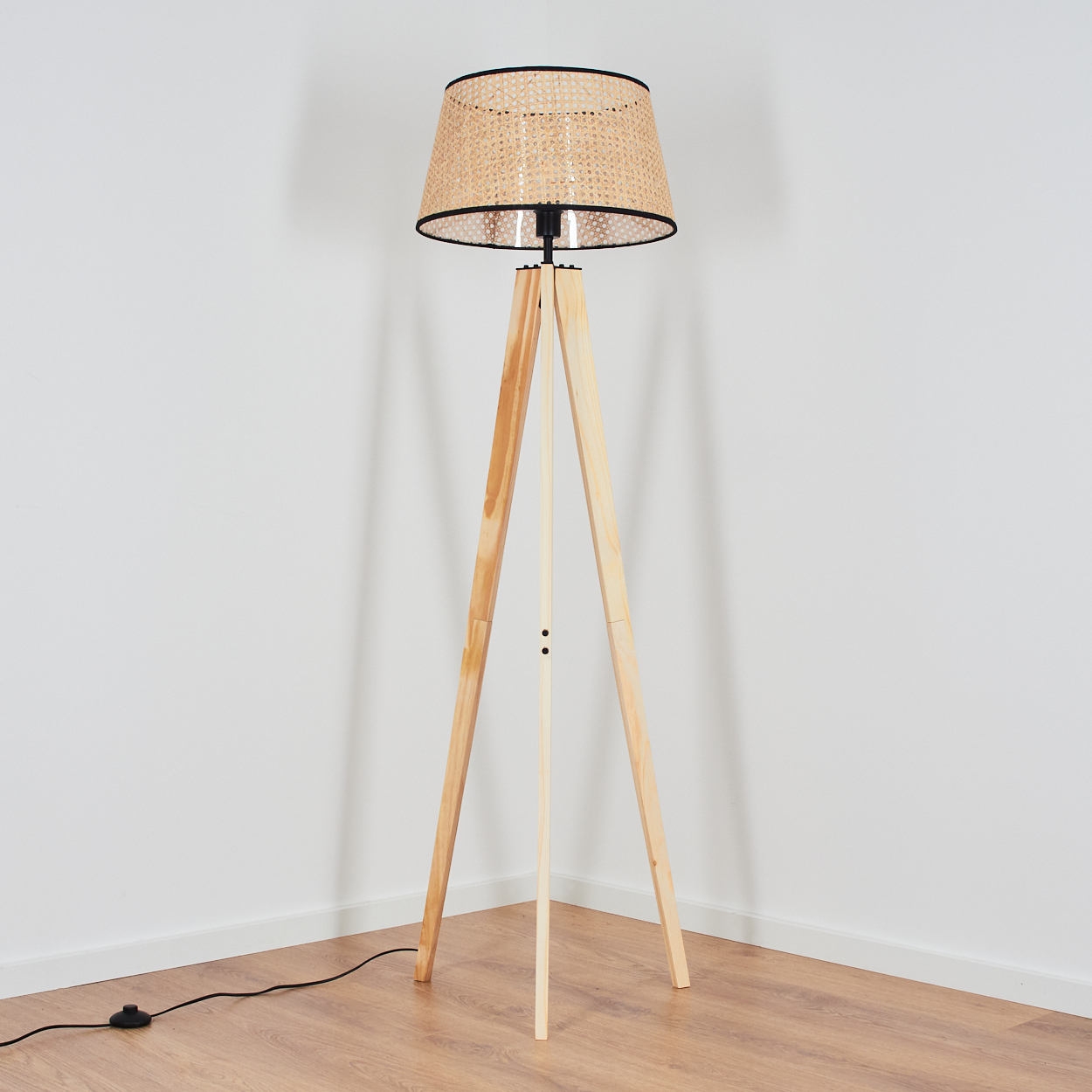 Eine hölzerne Stativ-Stehlampe mit gewebtem Lampenschirm steht in einer Ecke. Der Boden ist aus Holz, die Wände sind weiß.