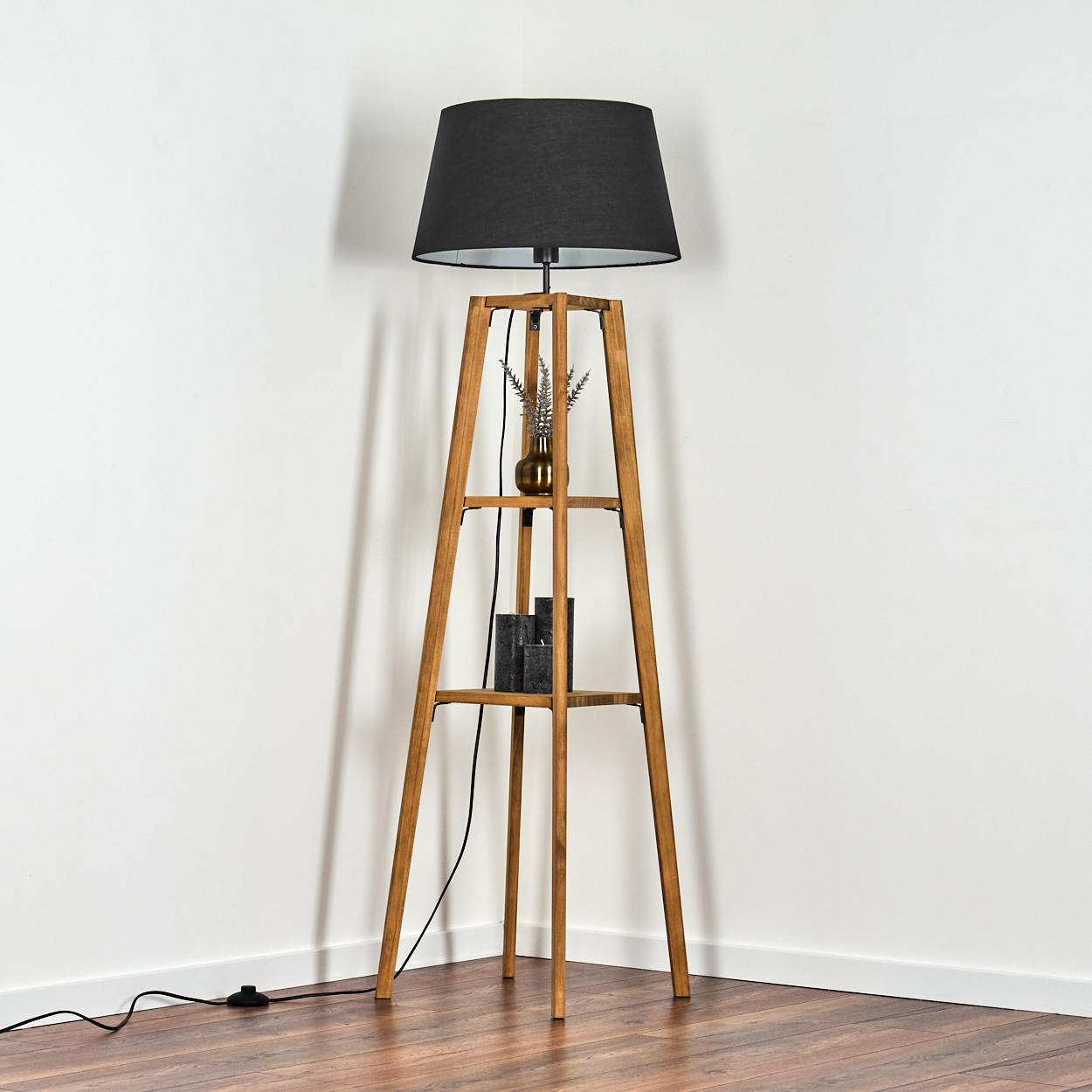 Eine Stehlampe mit schwarzem Lampenschirm steht in einer Ecke. Auf dem Regal stehen eine Vase mit Pflanzen und darunter Kerzen.