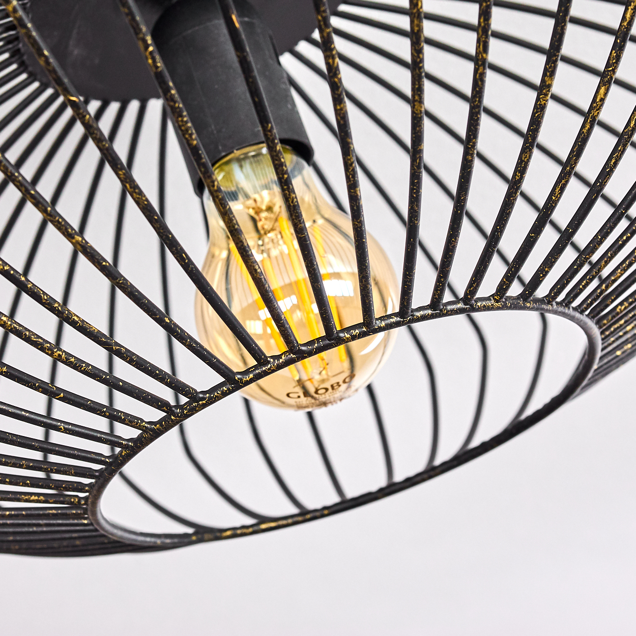 Nahaufnahme einer schwarzen Drahtgitterlampe mit einem warm getönten Leuchtmittel. Der Metallrahmen hat goldene Akzente auf weißem Hintergrund.