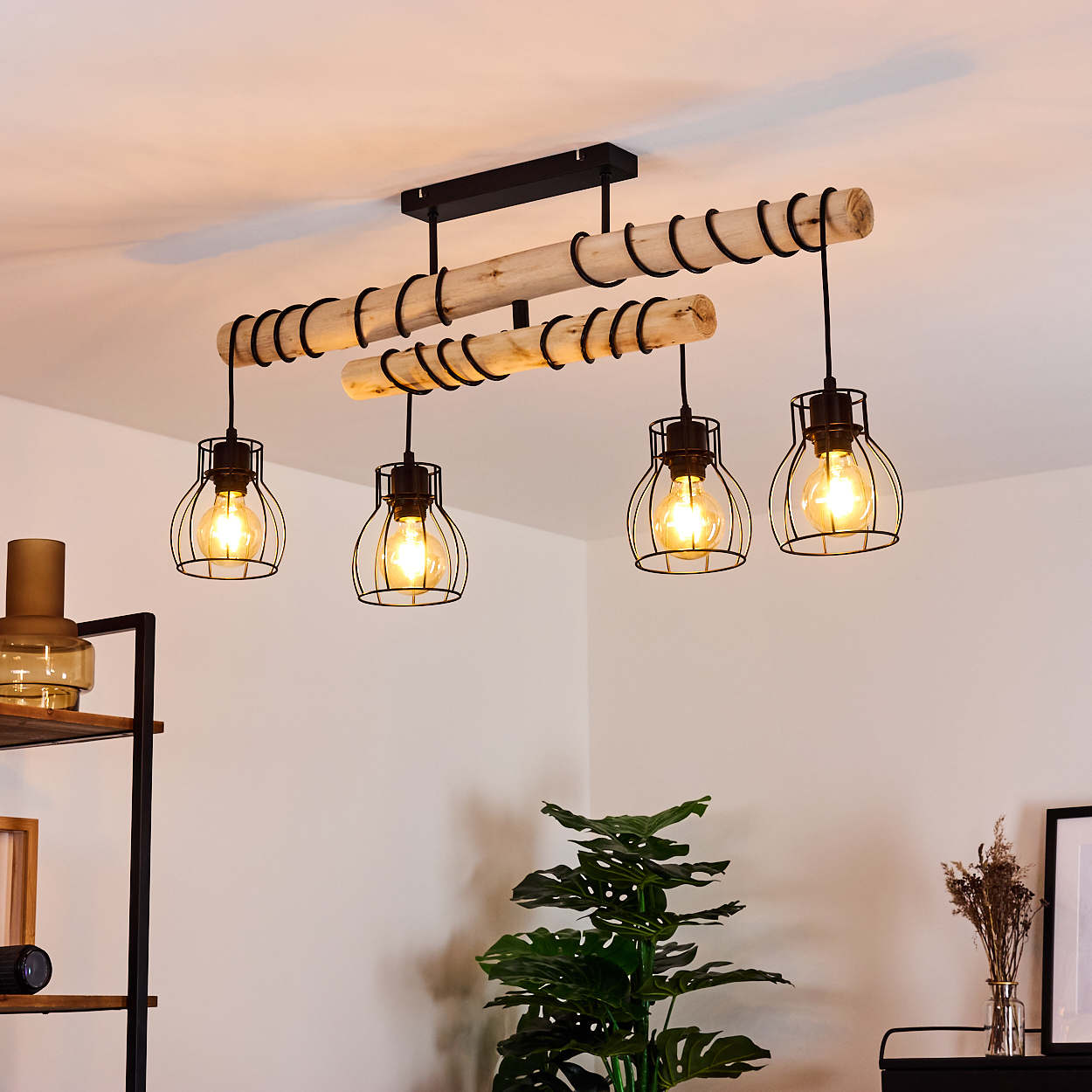 Ein Kronleuchter aus Holzbalken mit vier Lampen hängt von der Decke. Eine Pflanze und ein Regal sind im Raum sichtbar.