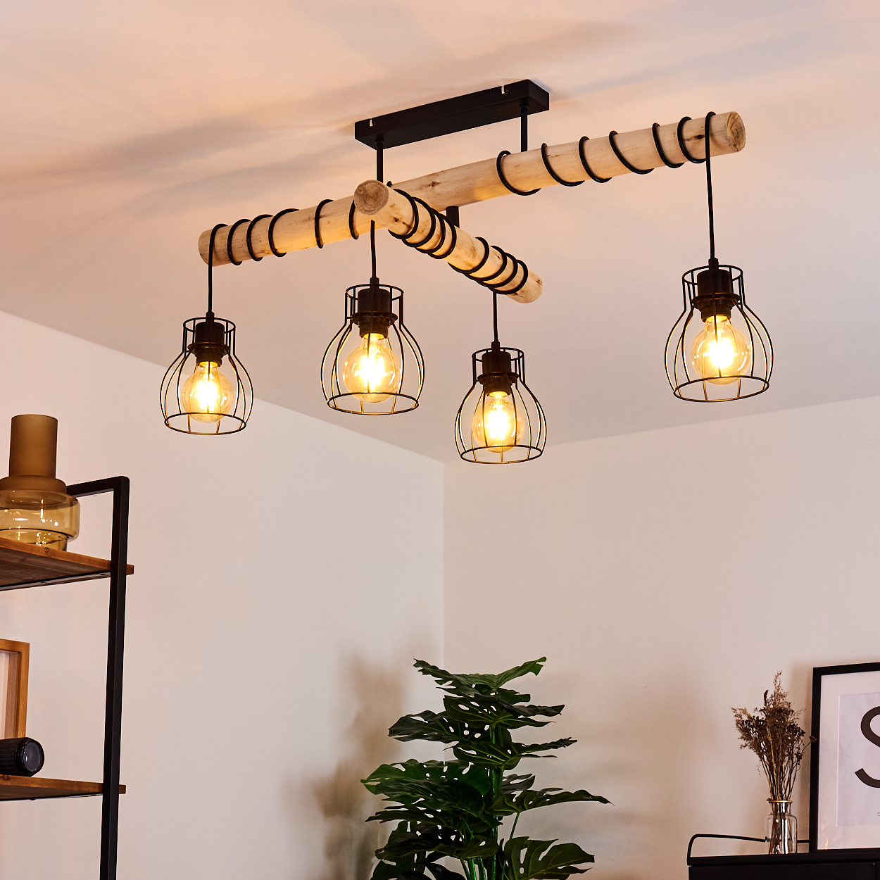 Ein Holzkronleuchter mit vier Käfiglampen hängt von einer weißen Decke. Darunter sind eine Pflanze und Dekoration zu sehen.