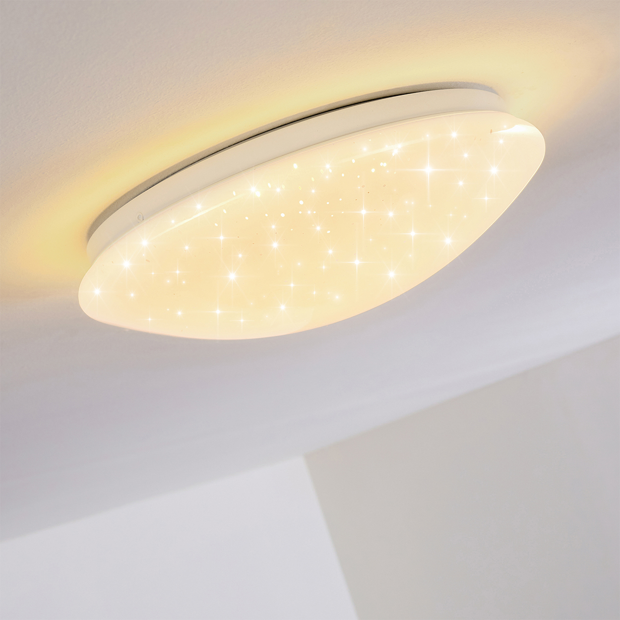 Eine weiße, ovale Deckenleuchte mit sternförmigem Muster. Das Licht ist an und strahlt warm. Die Decke ist weiß.
