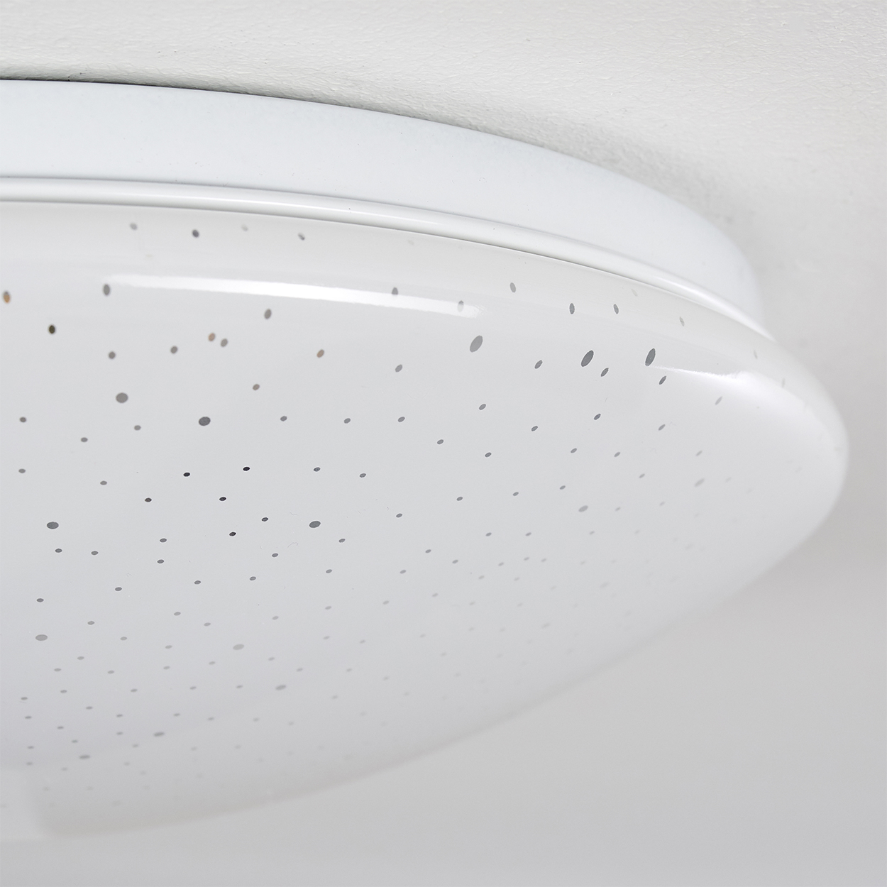 Weiße, runde Deckenleuchte mit kleinen dunklen Punkten, an einer weißen Decke montiert. Das Licht ist teilweise sichtbar.