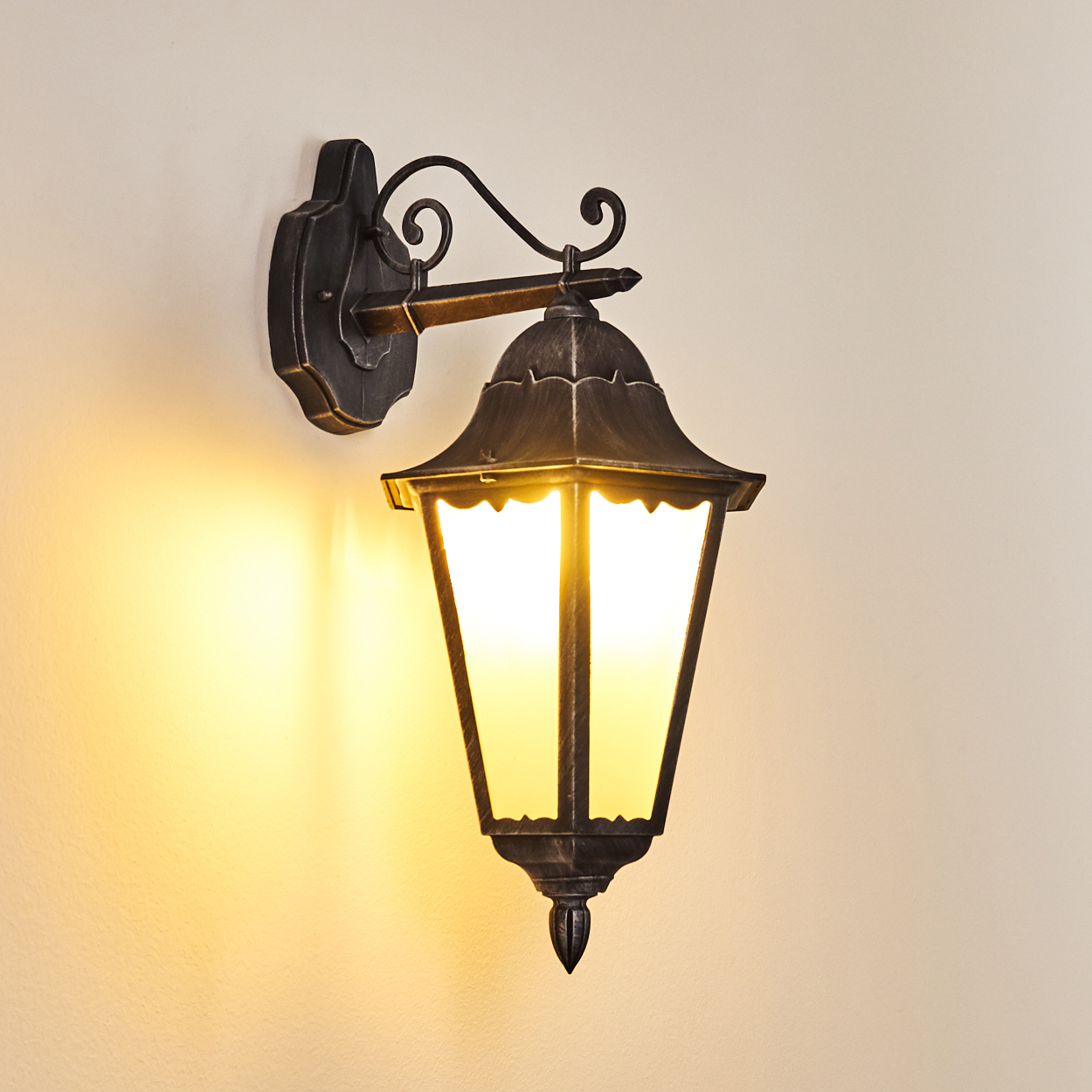 Eine an der Wand montierte, verzierte schwarze Lampe mit einem beleuchteten, rechteckigen Glaskasten. Die Lampe wirft einen warmen gelben Schein auf die Wand.