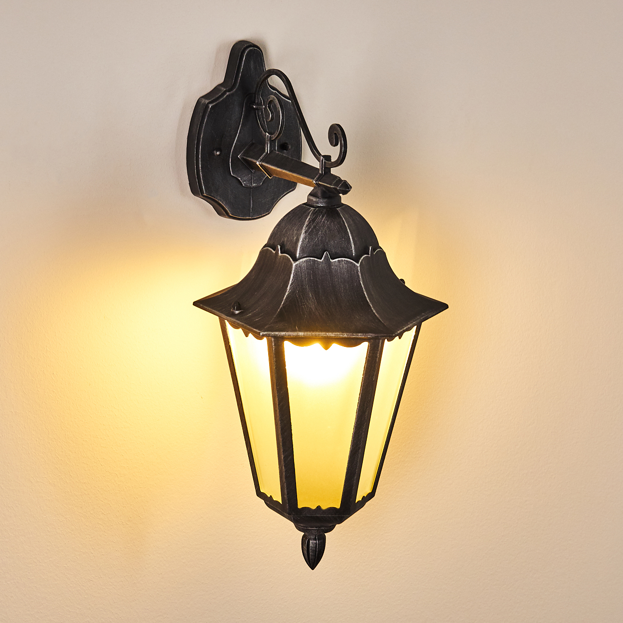 Eine Außenwandleuchte, schwarzes Metall mit goldgelbem Licht. Sie ist an einer beigefarbenen Wand mit dekorativem Design befestigt.