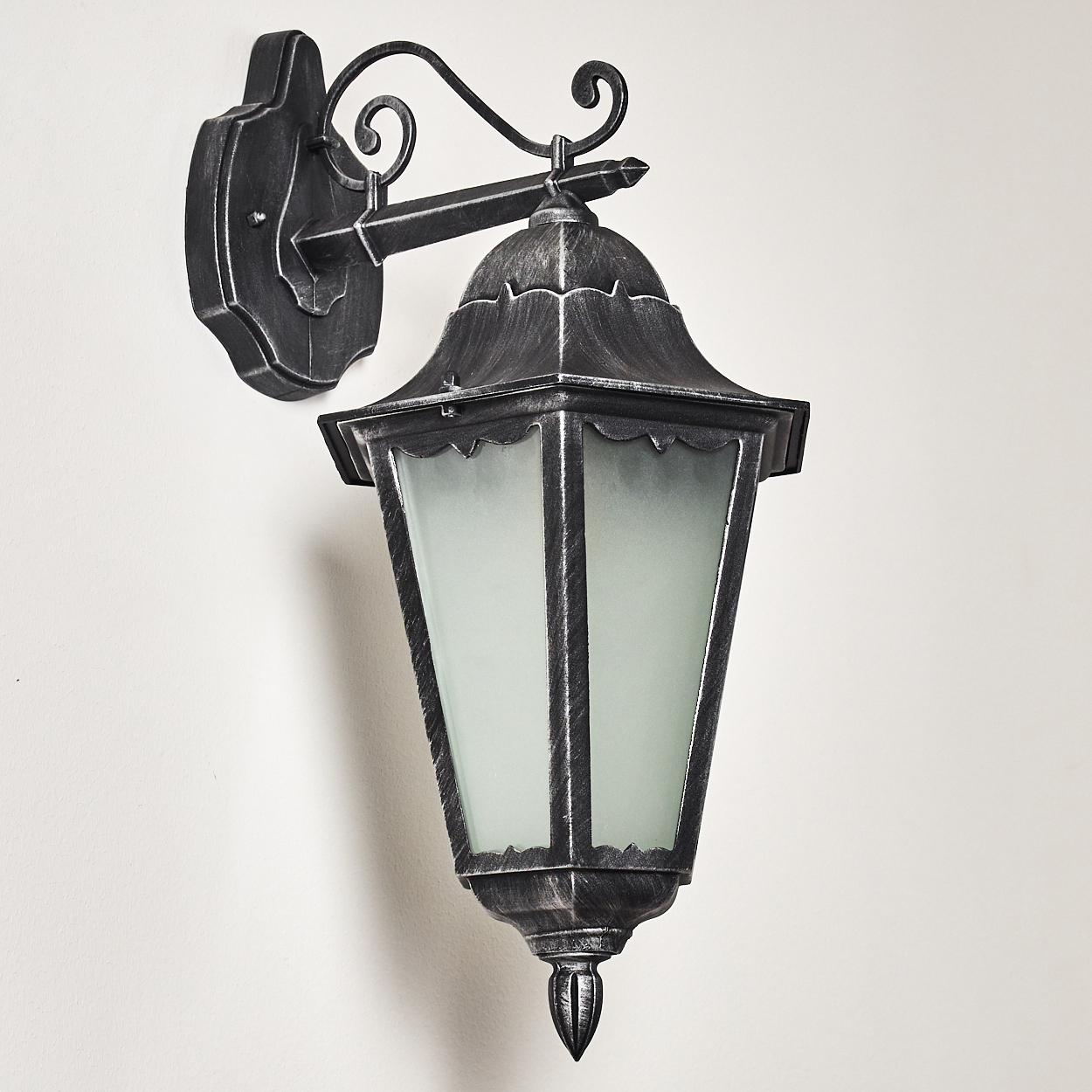 Eine dekorative, silberne Außenlampe an einer weißen Wand. Die Lampe hat einen Glasschirm und kunstvolles Metallwerk.