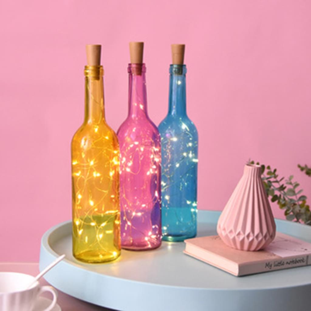 Drei bunte Glasflaschen mit Korken und Lichterketten. Sie stehen auf einem blauen Tablett mit einer rosa Vase und einem Buch.