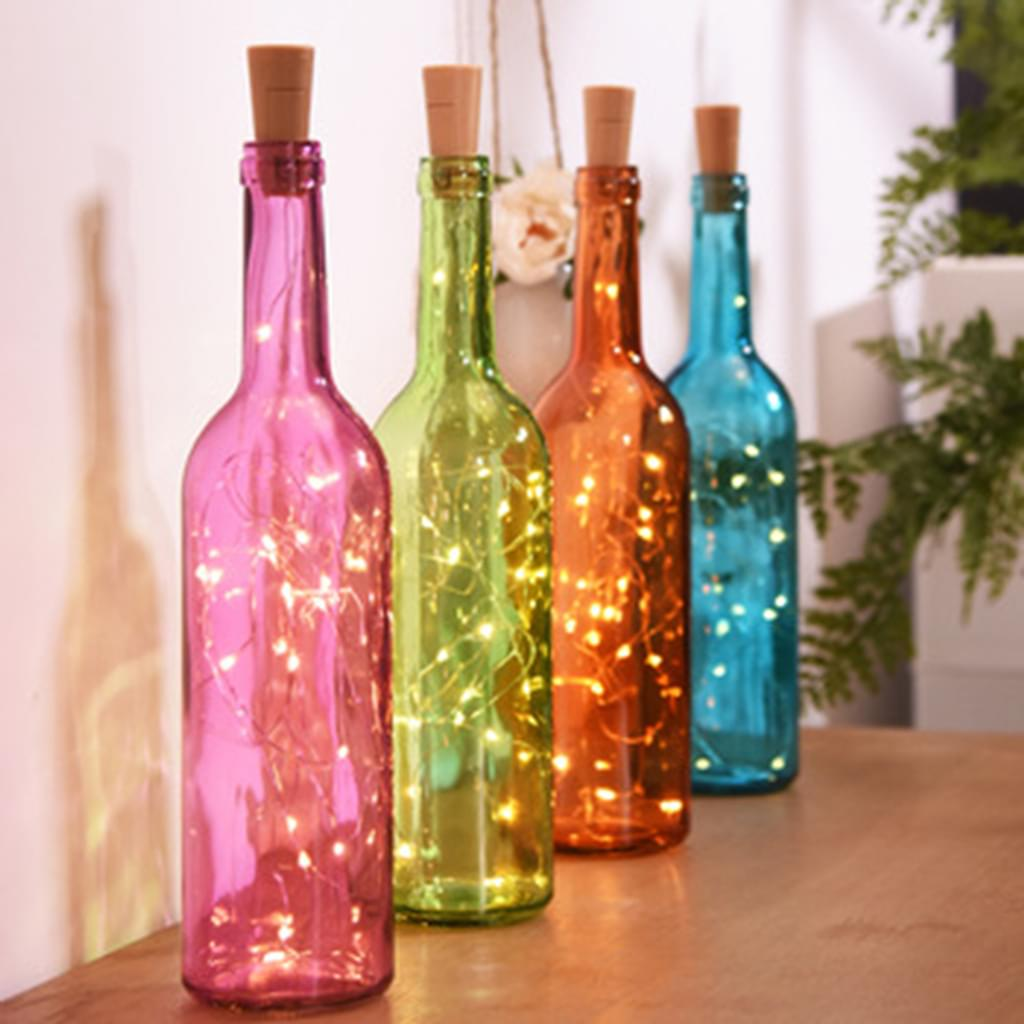 Vier farbige Glasflaschen mit Korken und Feenlichtern. Rosa, grüne, orange und blaue Flaschen stehen auf Holz.