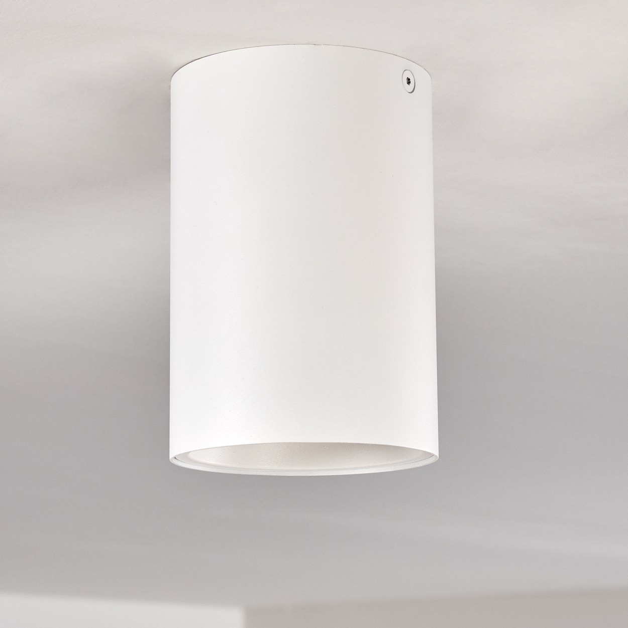 Eine weiße zylindrische Deckenleuchte. Sie hängt an einer weißen Decke. Das Licht hat eine kleine Schraube an der Seite.