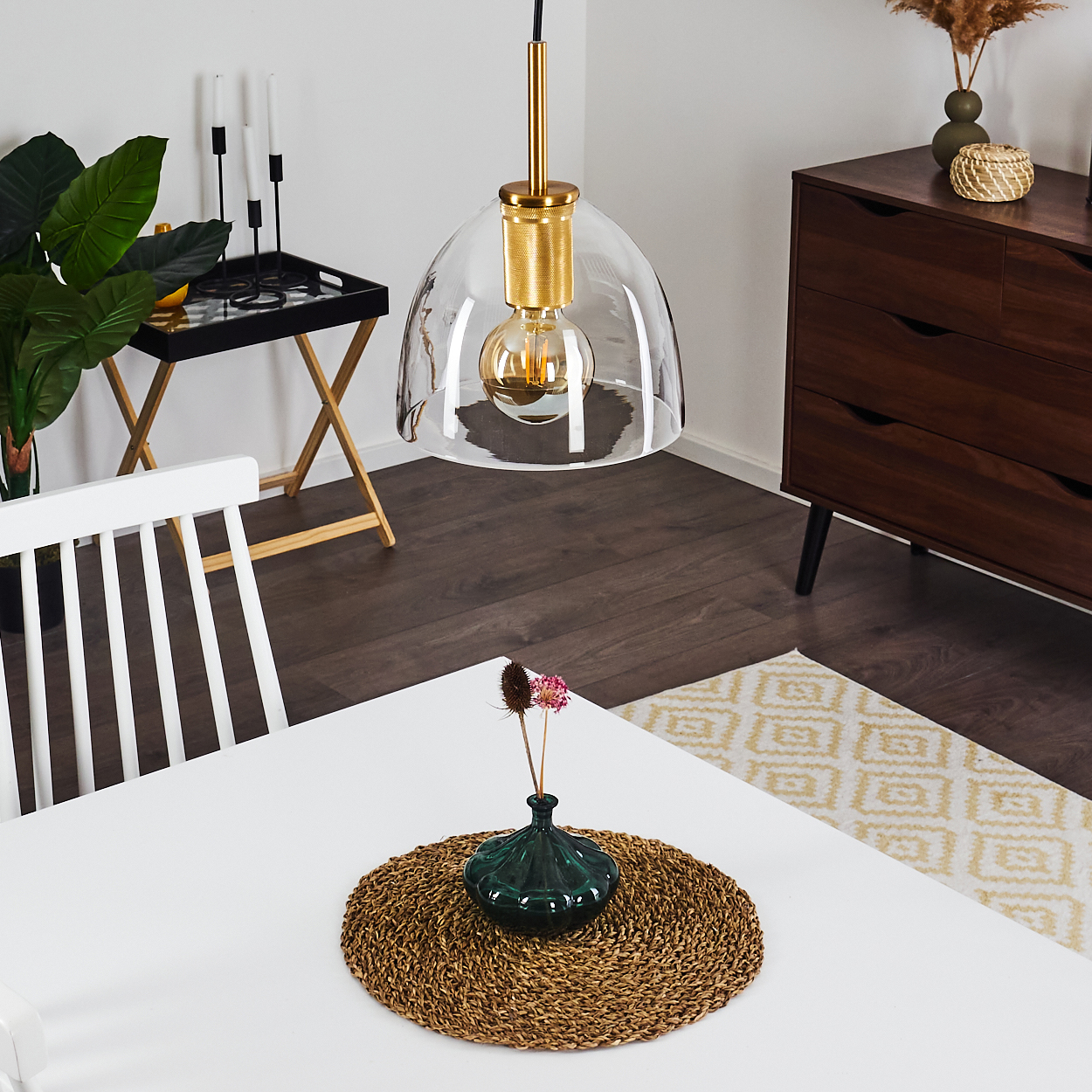 Ein Esszimmer mit einem weißen Tisch, einer Pendelleuchte, einer Vase mit Blumen und einem Holzschrank im Hintergrund.
