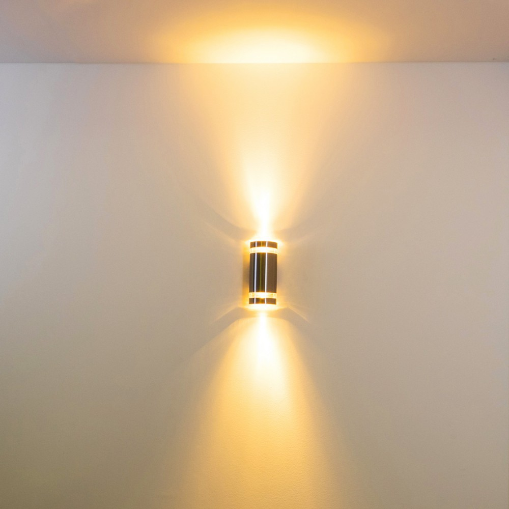 Eine Wandleuchte strahlt warmes Licht nach oben und unten aus. Die Leuchte ist schwarz und goldfarben und an einer weißen Wand angebracht.