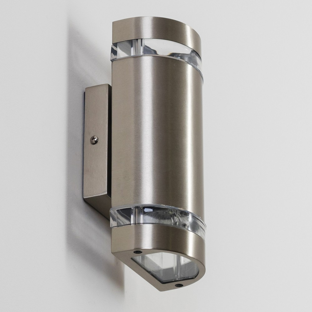 Eine metallische Wandleuchte ist an einer weißen Wand montiert. Die Lampe hat einen zylindrischen Körper mit klarer, gewölbter Ober- und Unterseite.