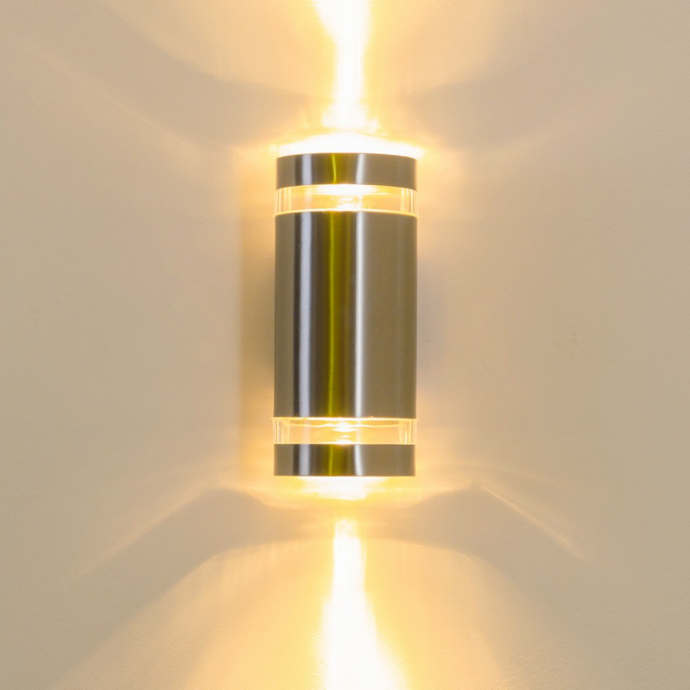 Eine silberne zylindrische Wandlampe mit transparenten Abschnitten, die hellgelbes Licht nach oben und unten an eine beige Wand abgibt.