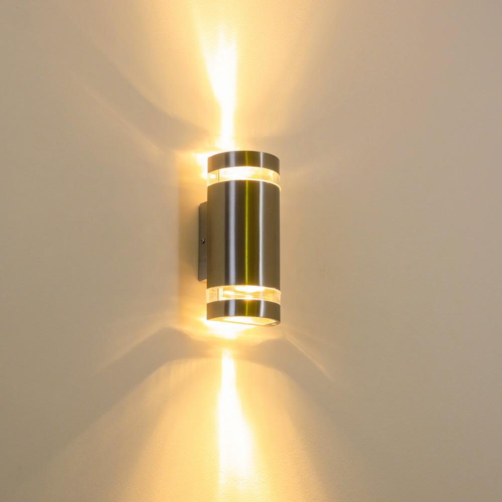 Eine zylindrische, silberne Außenwandleuchte mit hellem Licht, das nach oben und unten auf eine beige Wand scheint.