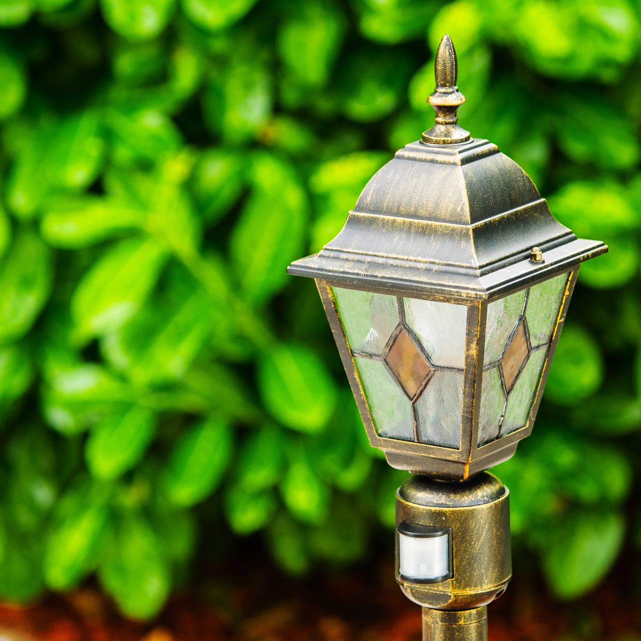 Eine dekorative Außenlampe mit grünem, rautenförmigem Glas und grünem, belaubtem Hintergrund.