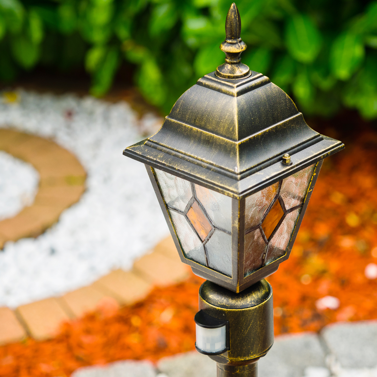 Eine verzierte Außenlampe mit Buntglas und einem Sensor. Im Hintergrund ein verschwommener Gartenweg mit orangem Mulch und grünem Laub.