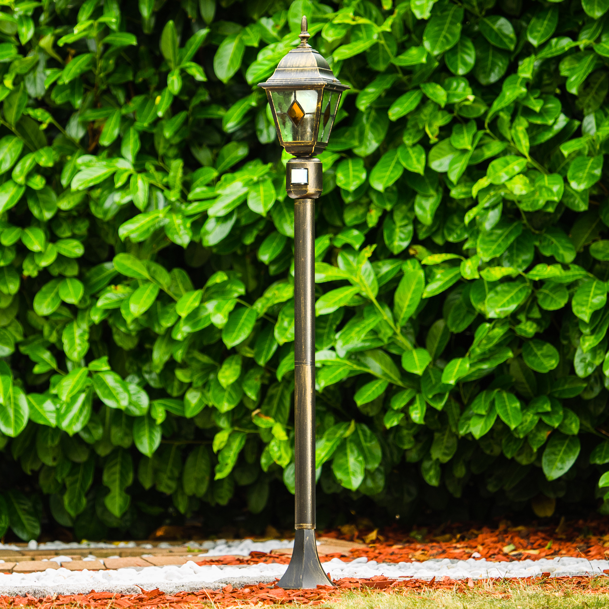 Eine verzierte Bronzelaterne steht vor einer dichten, grünen Hecke. Die Lampe ist beleuchtet. Weiße Steine und roter Mulch umgeben den Sockel.