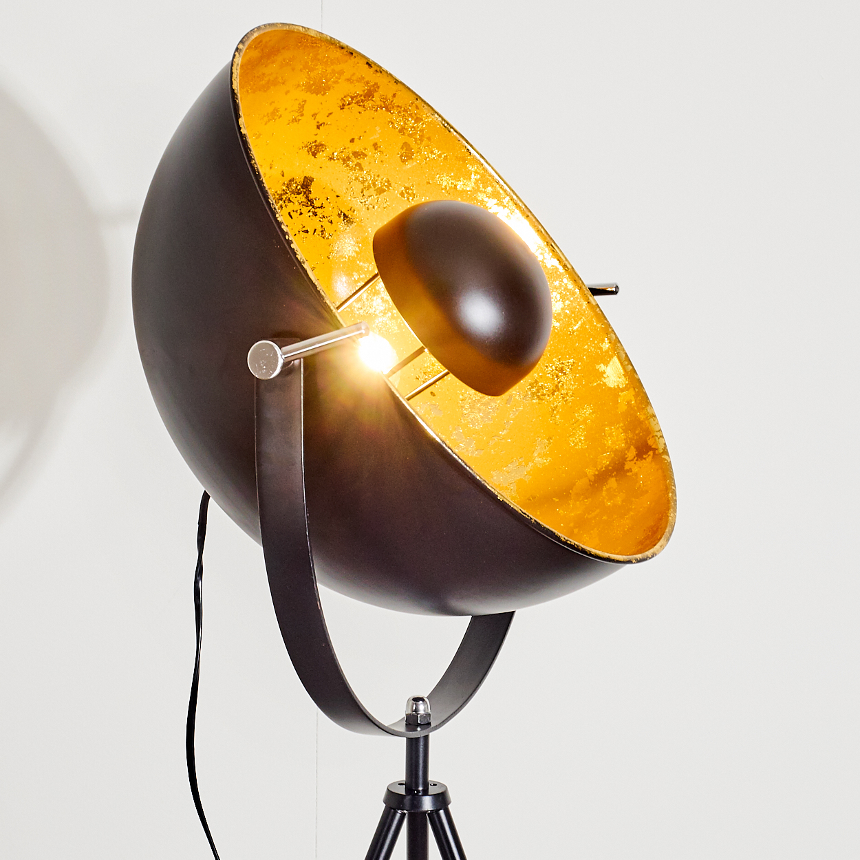 Eine schwarz-goldene Stehlampe. Die Lampe hat einen großen, schalenförmigen Kopf mit goldener Innenseite und schwarzer Außenseite, auf einem Stativ.