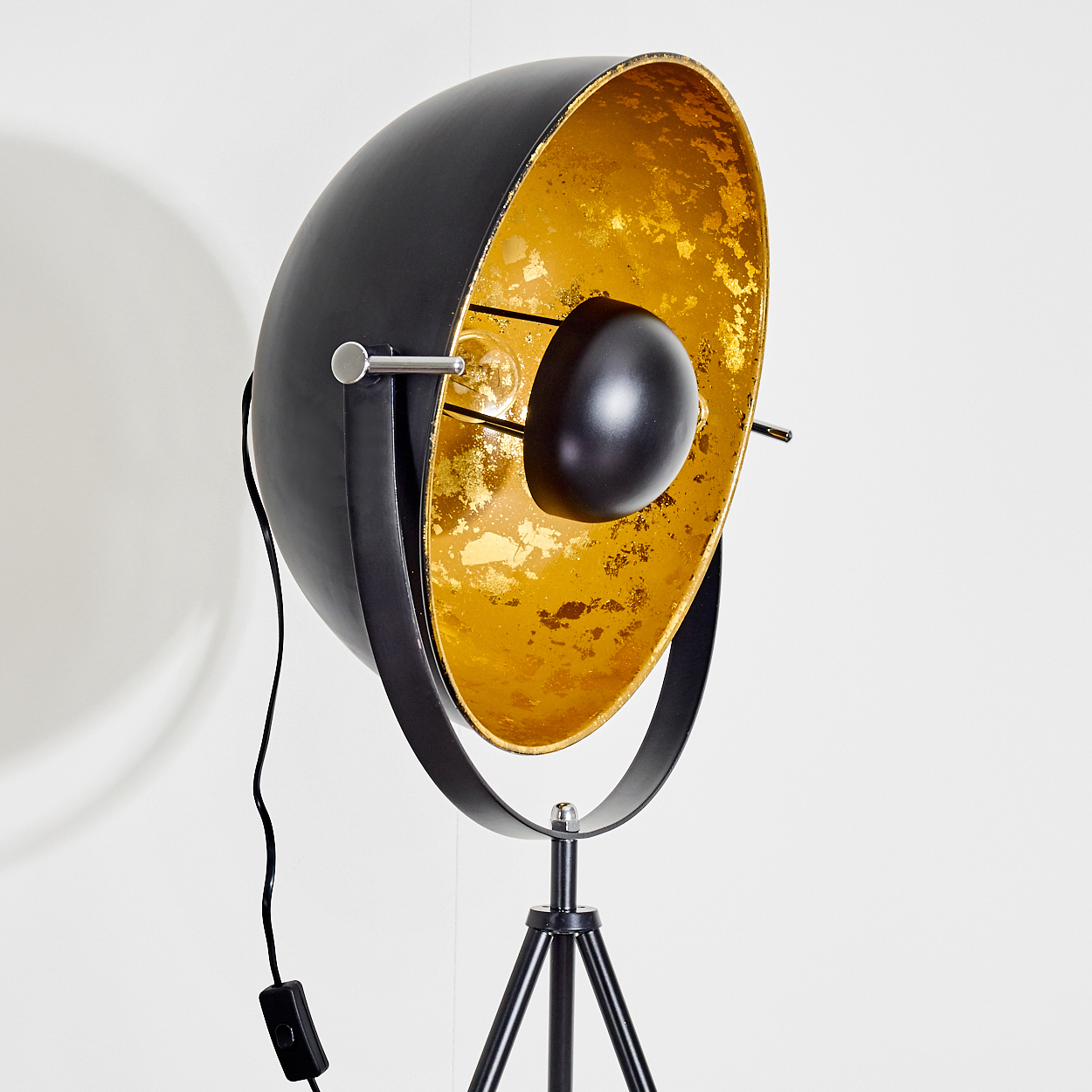 Eine schwarz-goldene Stehlampe. Die Lampe hat einen großen, runden Kopf mit goldener Innenseite. Sie steht auf einem schwarzen Stativfuß.