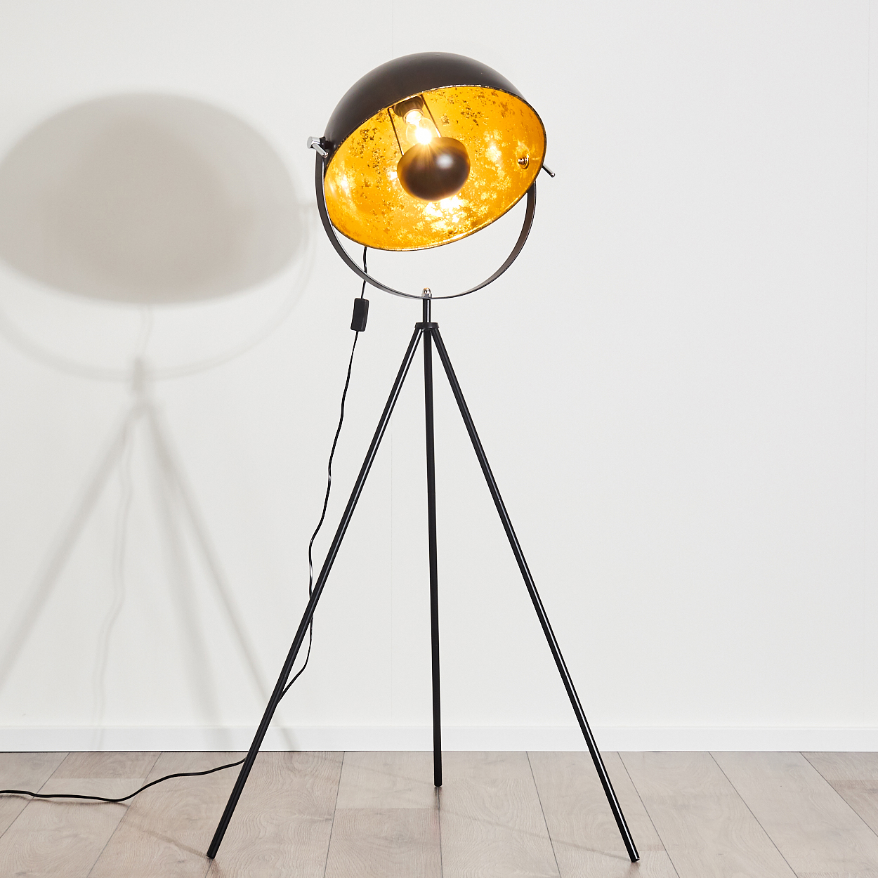 Eine schwarz-goldene Stehlampe steht auf einem Holzboden. Die Lampe wirft einen Schatten auf die weiße Wand. Die Lampe ist eingeschaltet.